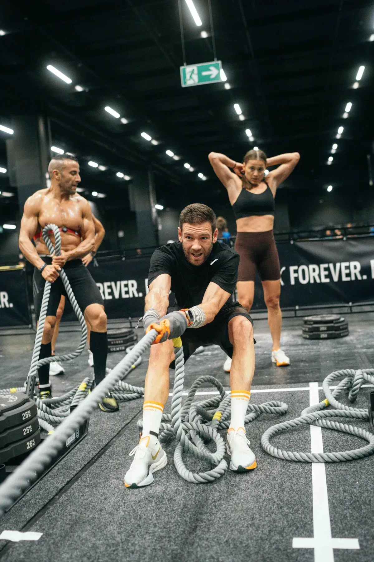 Three people doing rope exercises in a gym. Man pulls rope forward; others stand, one with arms behind head.