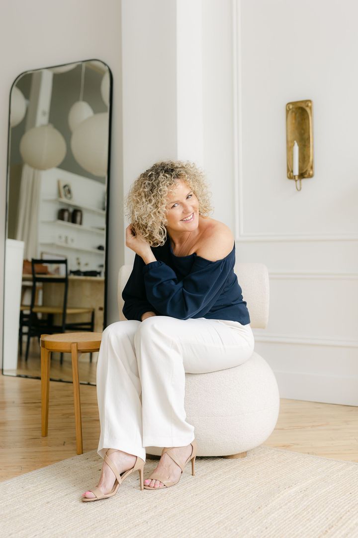 Woman with curly blonde hair, navy top, white pants, smiling in a bright room; seated on a round, off-white chair.