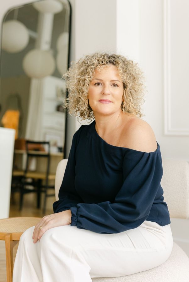 Woman with curly blonde hair, navy top, and white pants, sitting in a bright room.