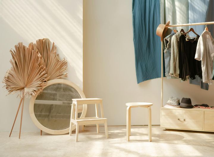 A light-filled room with clothes rack, stool, mirror, dried palm leaves, and a cabinet.