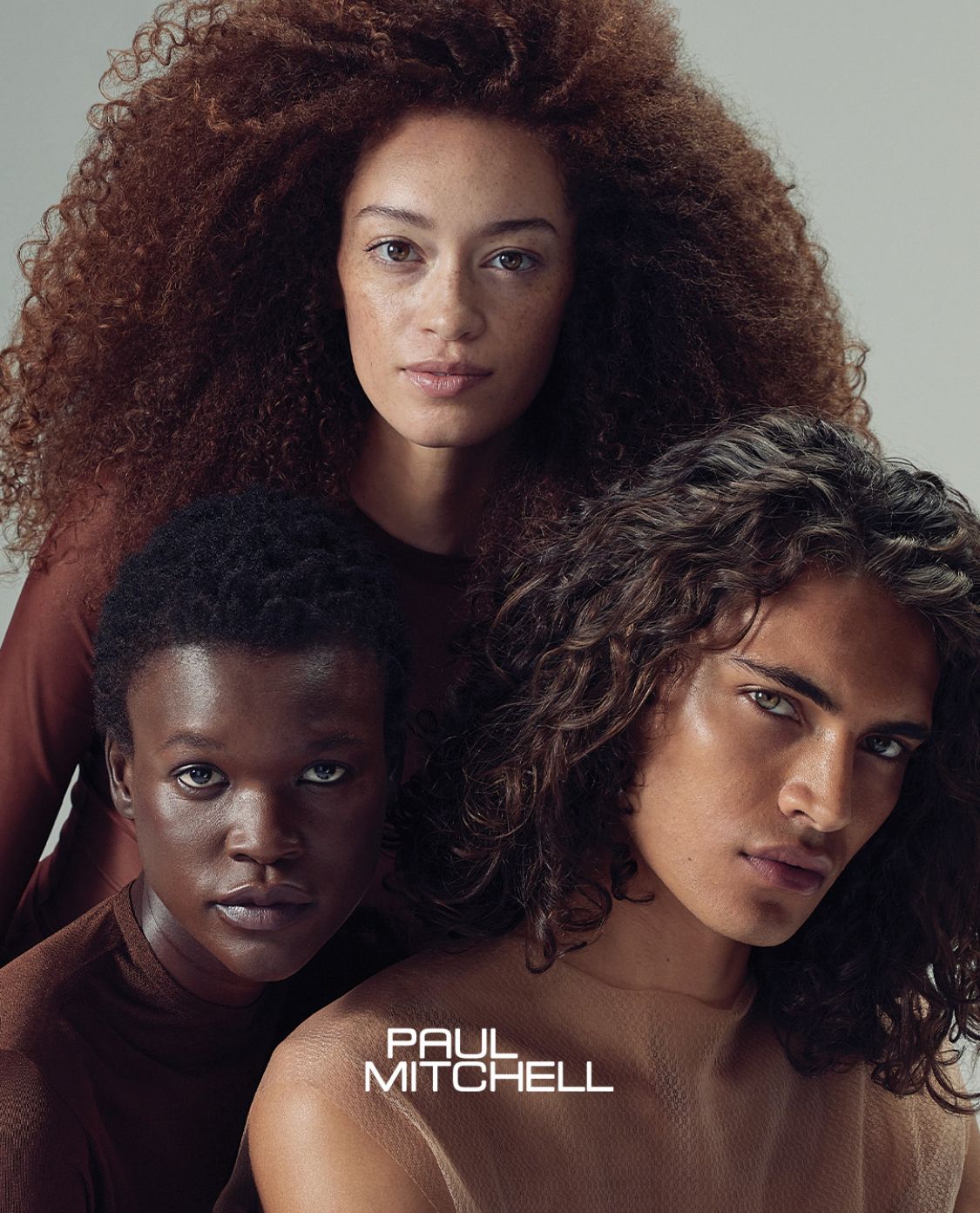 Tre persone con i capelli mossi posano su uno sfondo grigio sopra il logo di Paul Mitchell.