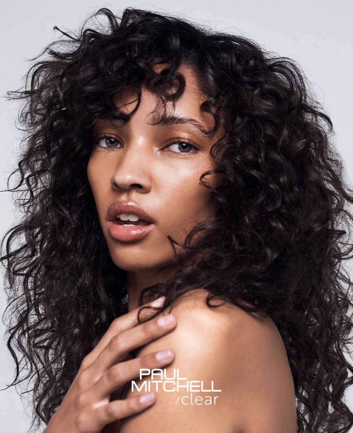 Una persona con una folta chioma scura e riccia guarda verso la telecamera, con il logo di Paul Mitchell visibile in basso.