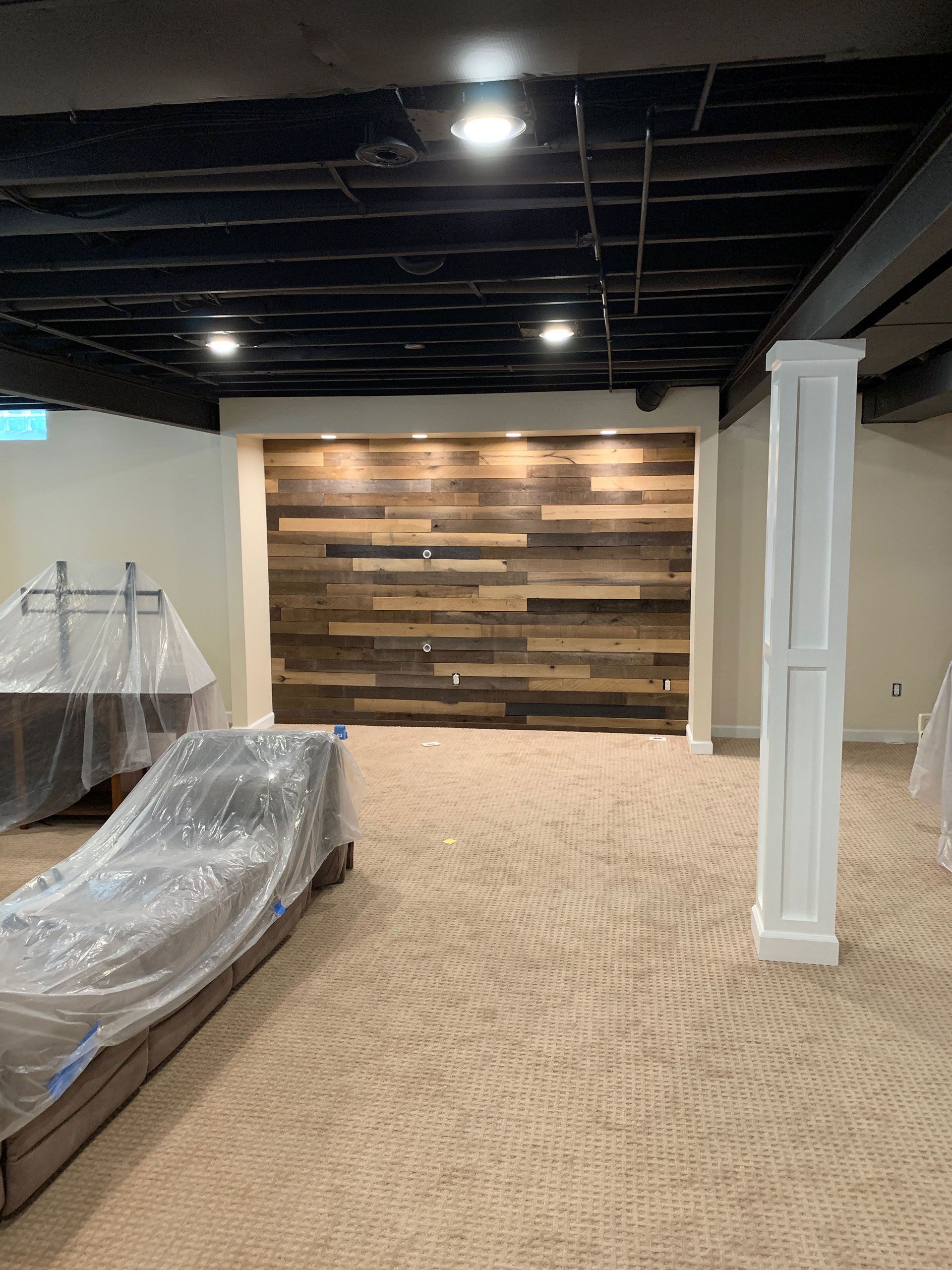 Basement with beige carpet, a dark wood-plank accent wall, a white pillar, and furniture covered in plastic sheeting.