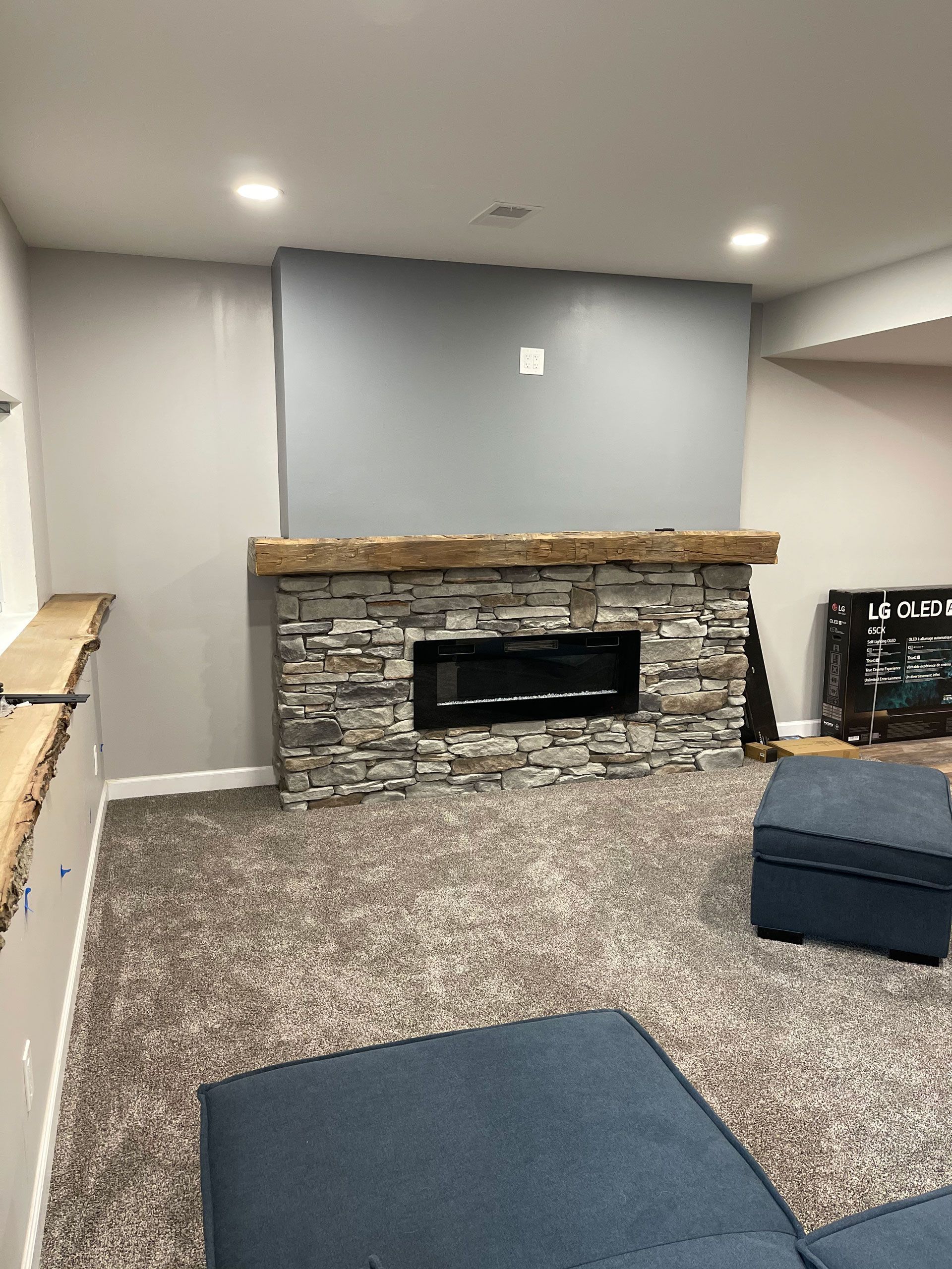 A basement living area featuring a stone fireplace with a rustic wood mantel, gray walls, and blue seating.