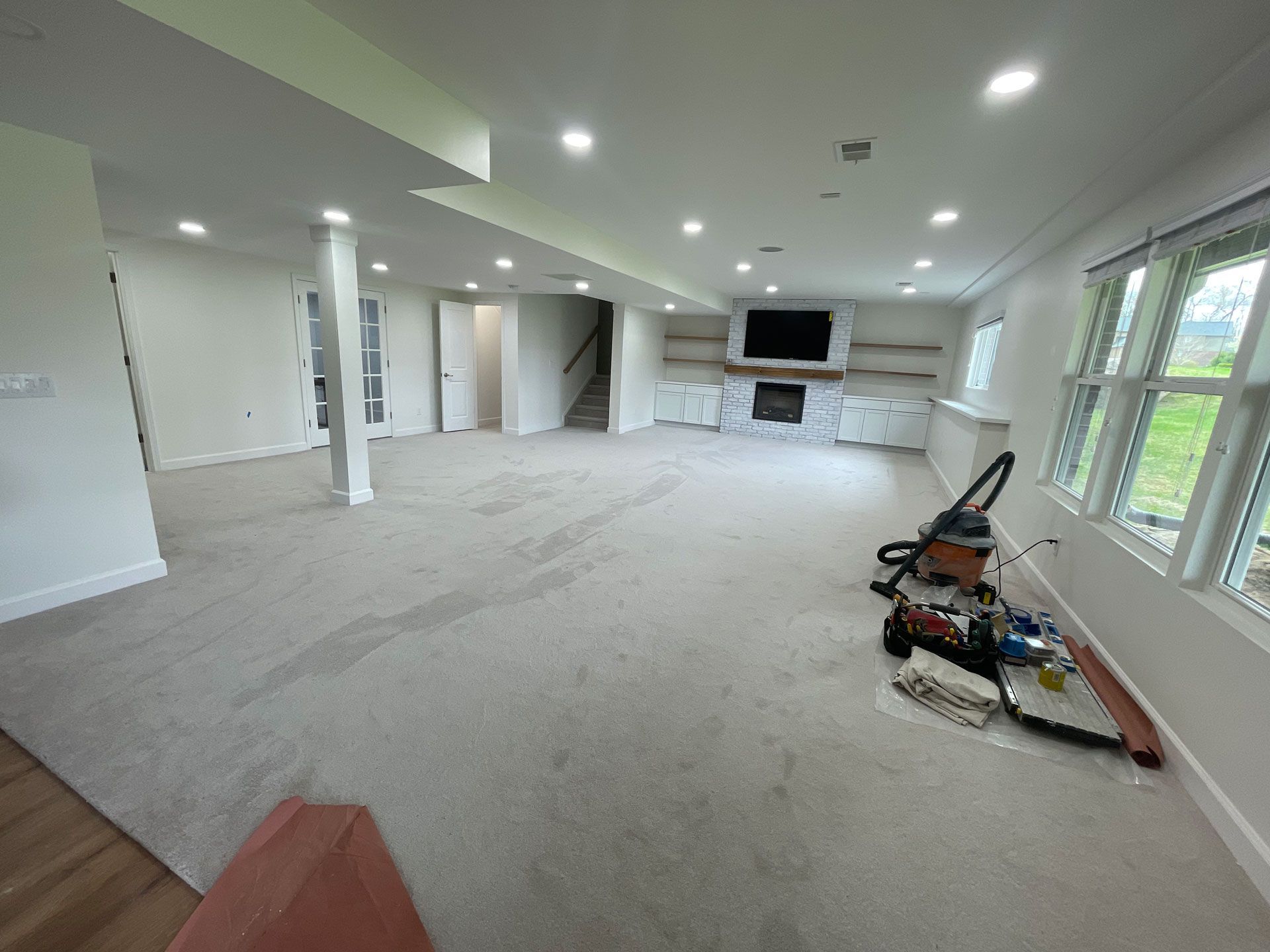 A spacious basement with gray carpet, recessed lighting, a stone fireplace, built-in shelving, and construction equipment.