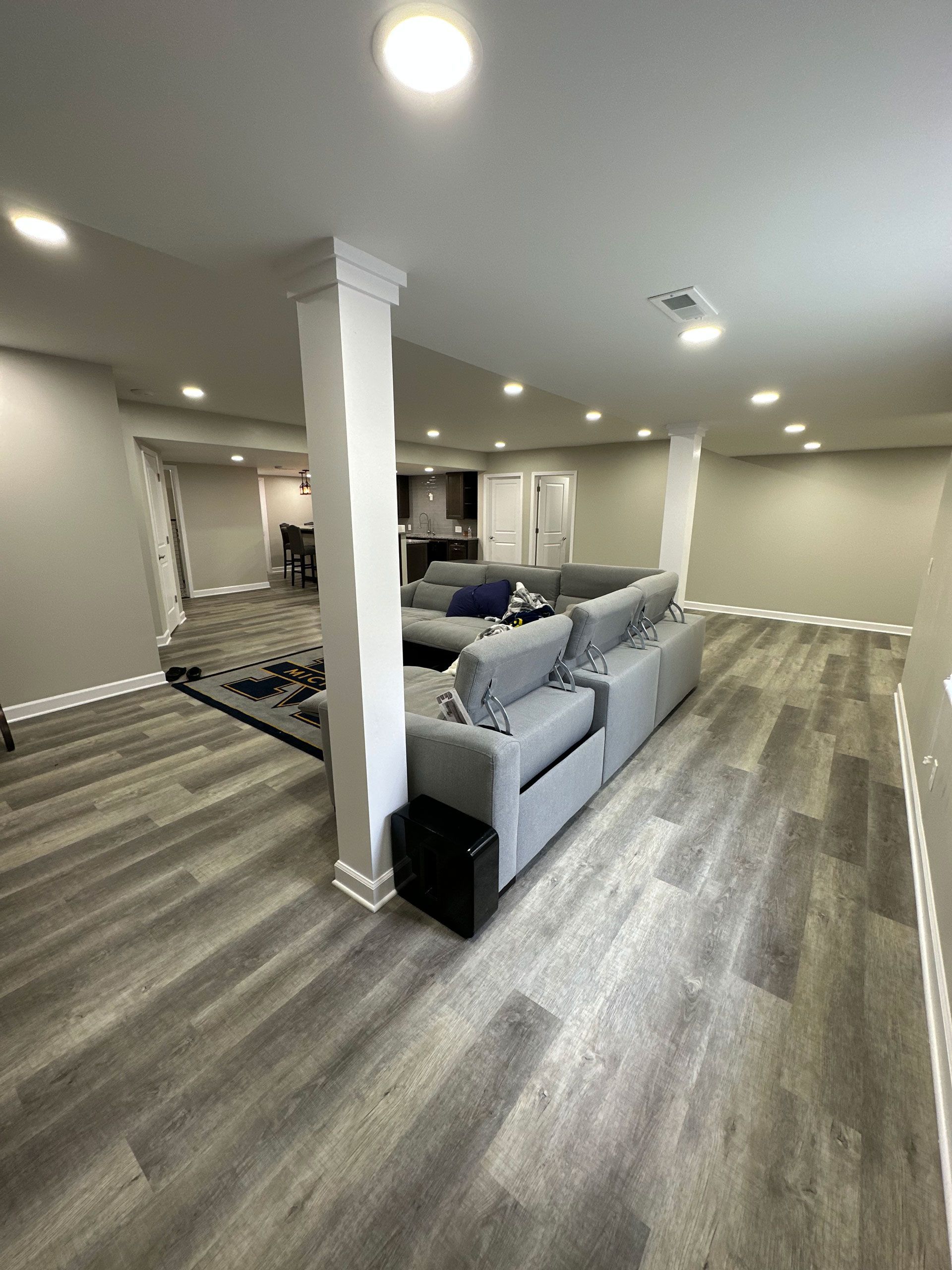 A finished basement with grey wood-look flooring, light grey walls, recessed lighting, and a grey sectional sofa.