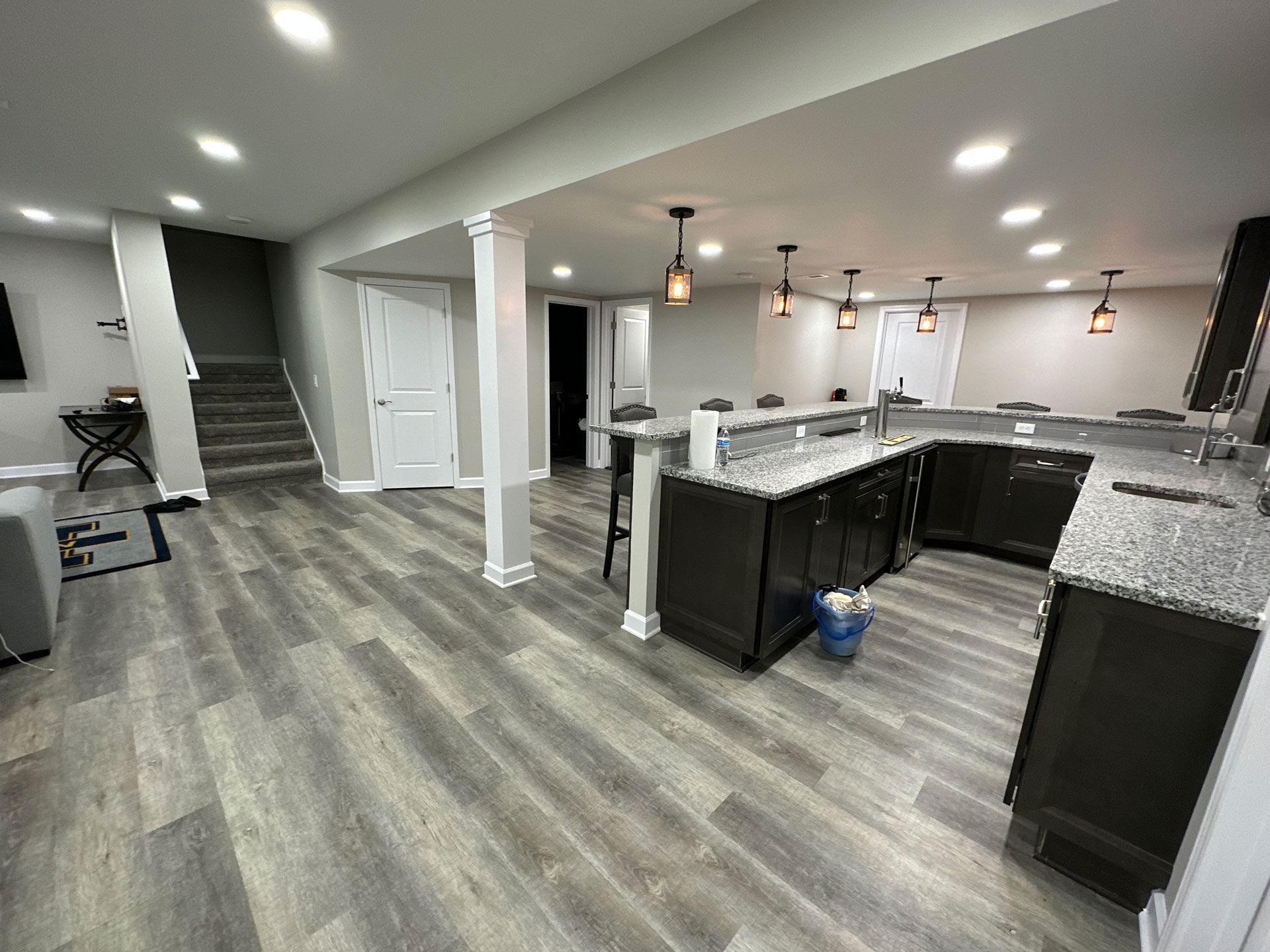A basement interior with gray wood-look flooring, a kitchen area featuring dark cabinets, granite counters, and pendant lights.