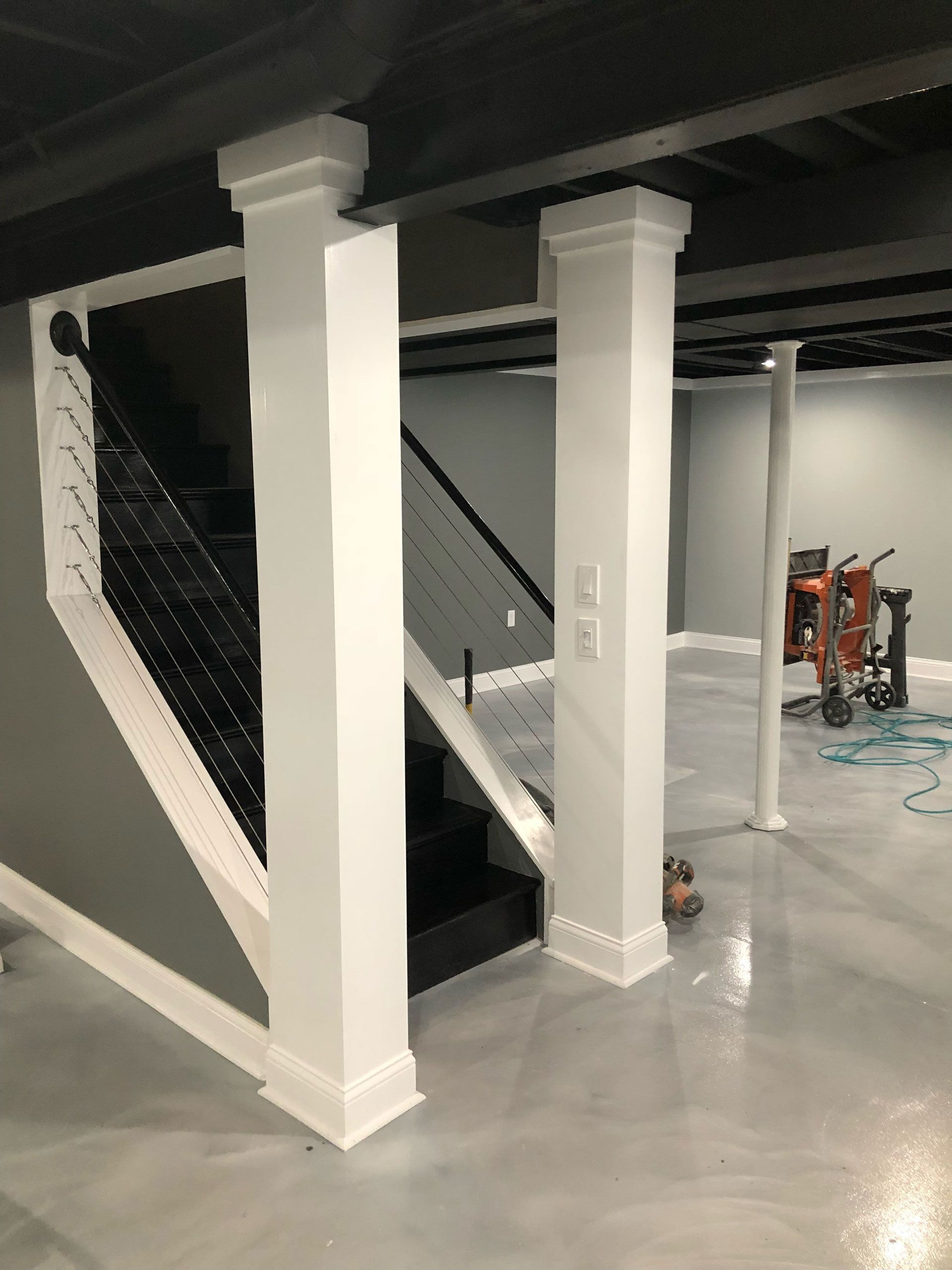 A view of a basement staircase with a modern cable railing, featuring two white pillars and a glossy grey floor.