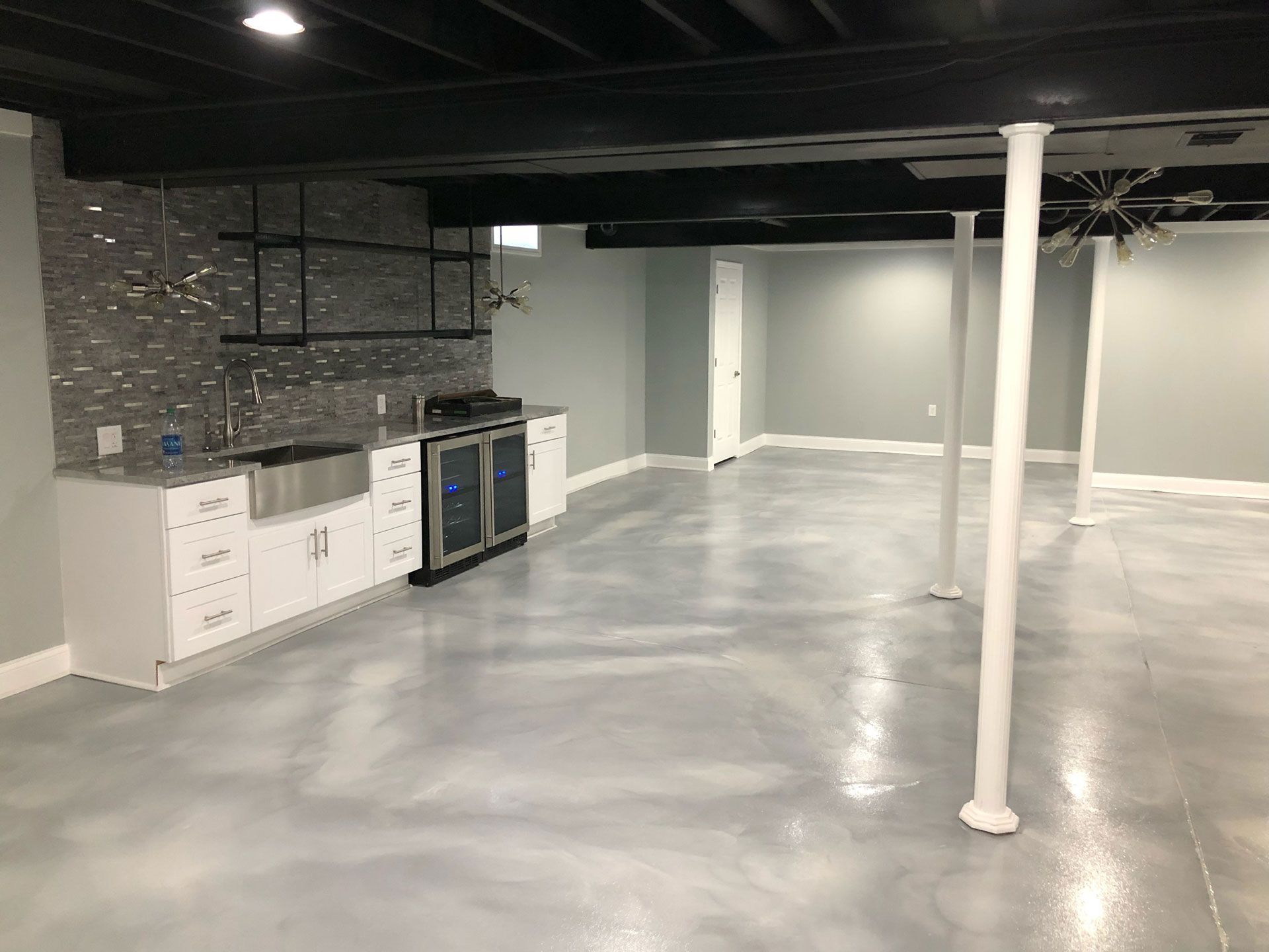 A basement kitchenette with white cabinets, a stone backsplash, and a metallic epoxy floor beneath black exposed beams.