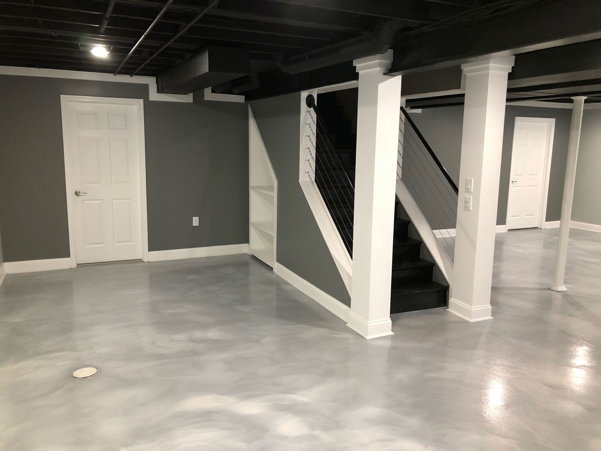 Modern basement with gray walls, white columns and trim, black exposed ceiling, and glossy gray concrete flooring.