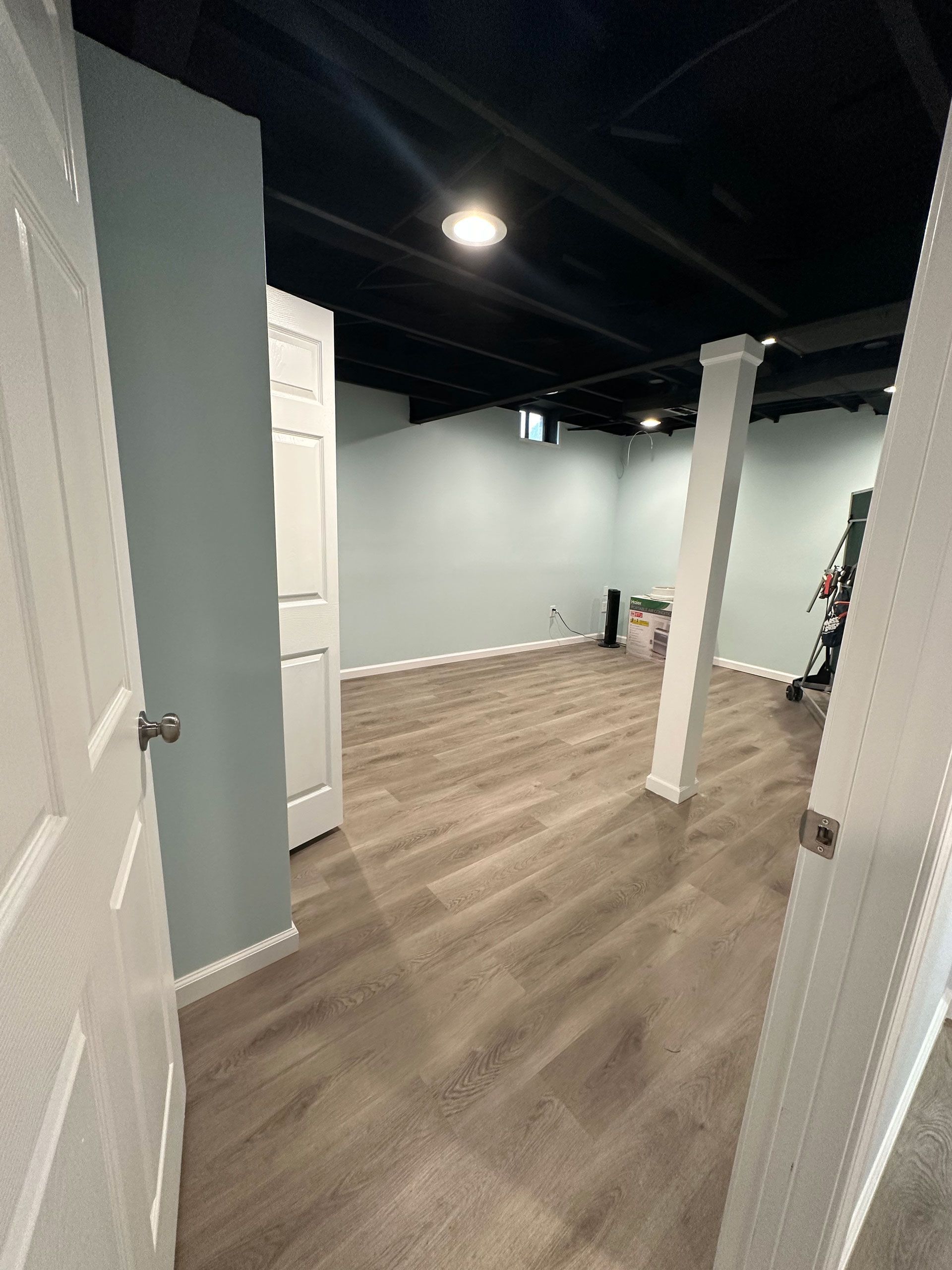 A basement room with light blue walls, wood-look flooring, a black painted ceiling, and a central white support column.
