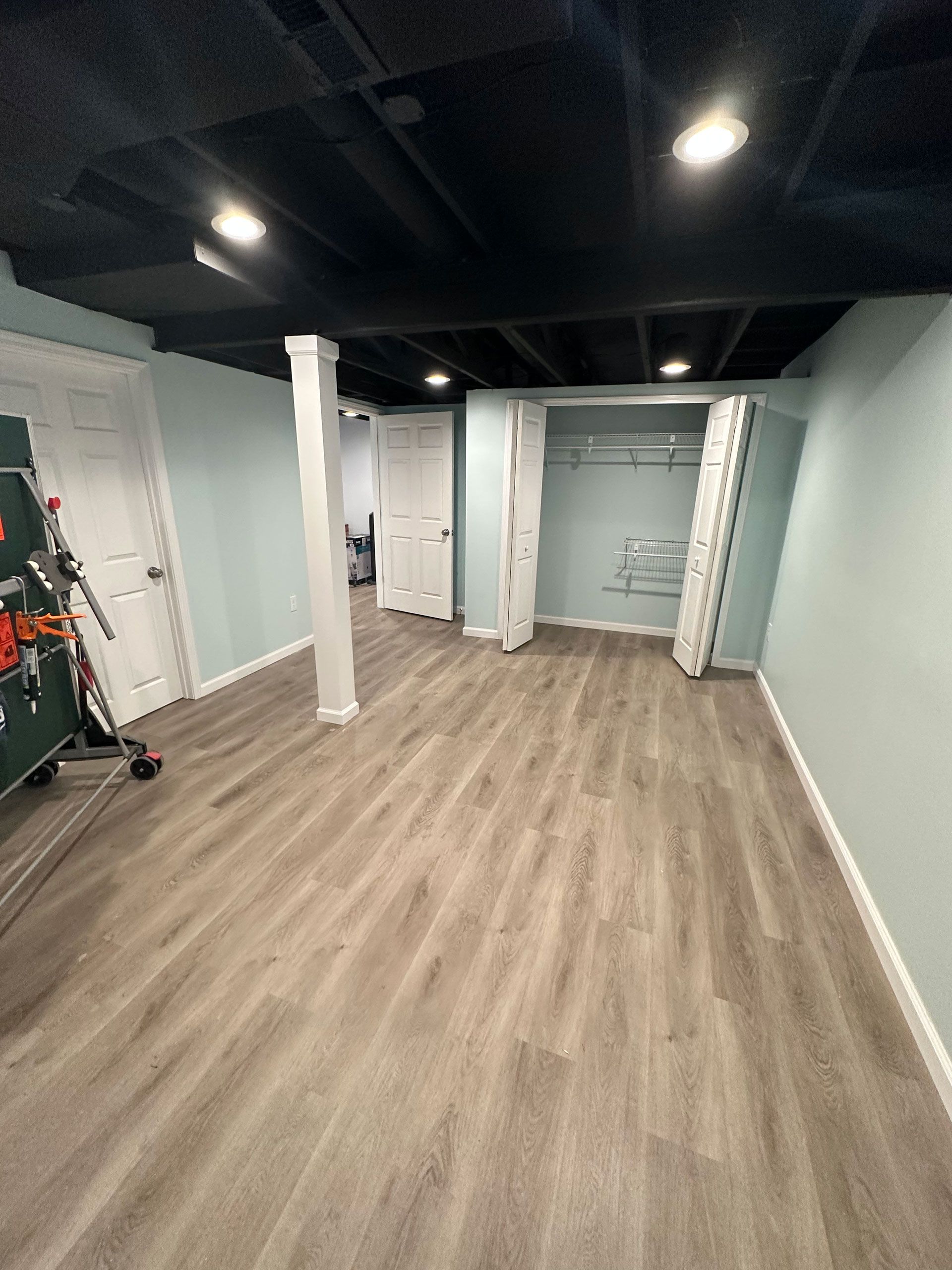 A basement room with light blue walls, gray wood-look flooring, a black exposed ceiling, and two white closet doors.