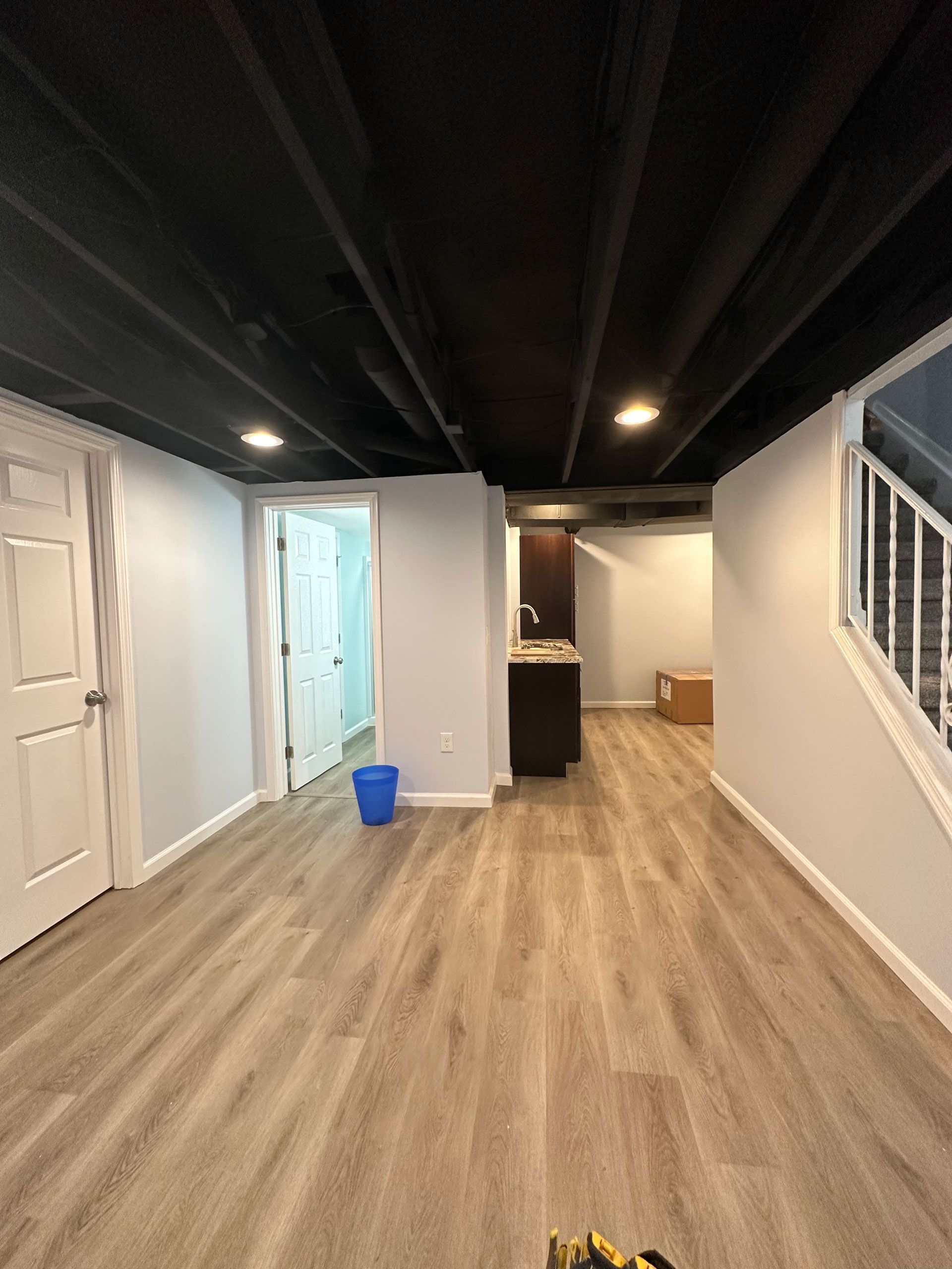 A basement with light gray walls, light wood-look flooring, a black-painted exposed ceiling, and a doorway to another room.