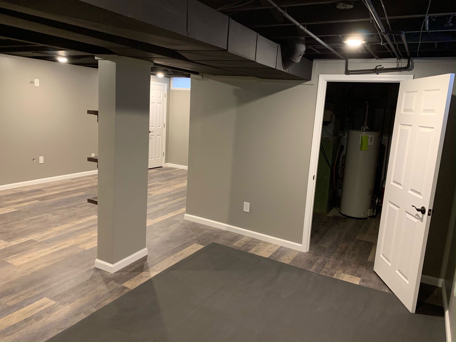 A basement room with gray wood-look flooring, light gray walls, a black ceiling, and an open utility closet door.