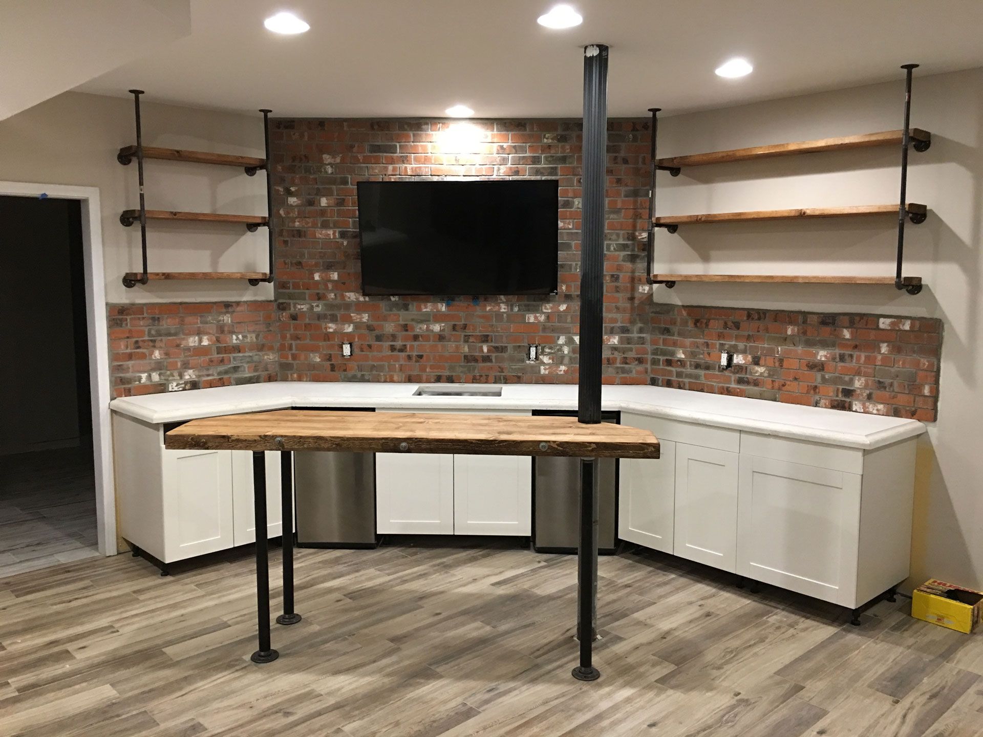 A modern basement bar with white cabinets, a live-edge wooden countertop, brick backsplash, and pipe-style wall shelving.