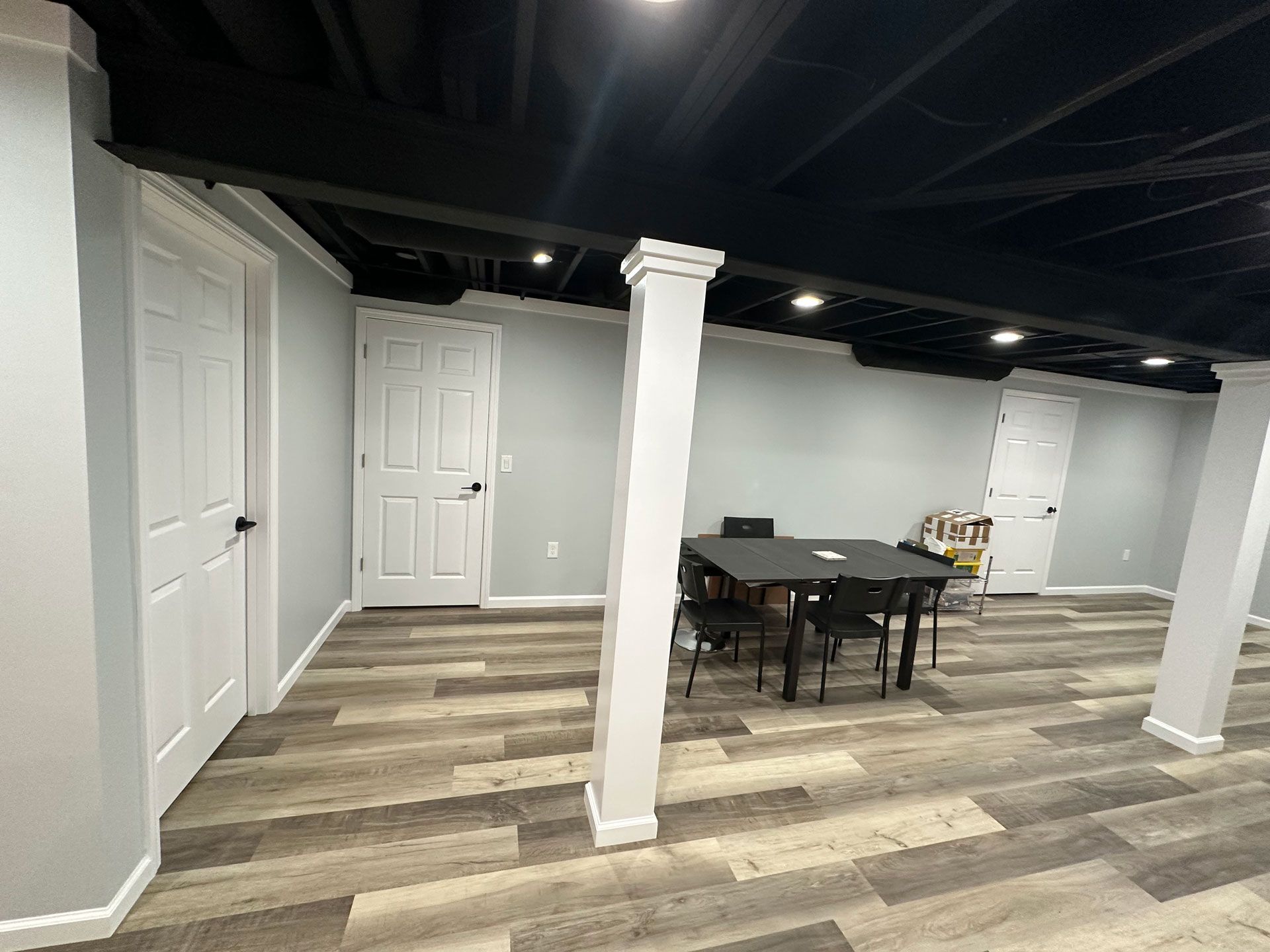 A basement interior with light gray walls, wood-look flooring, white support columns, and a table with chairs.