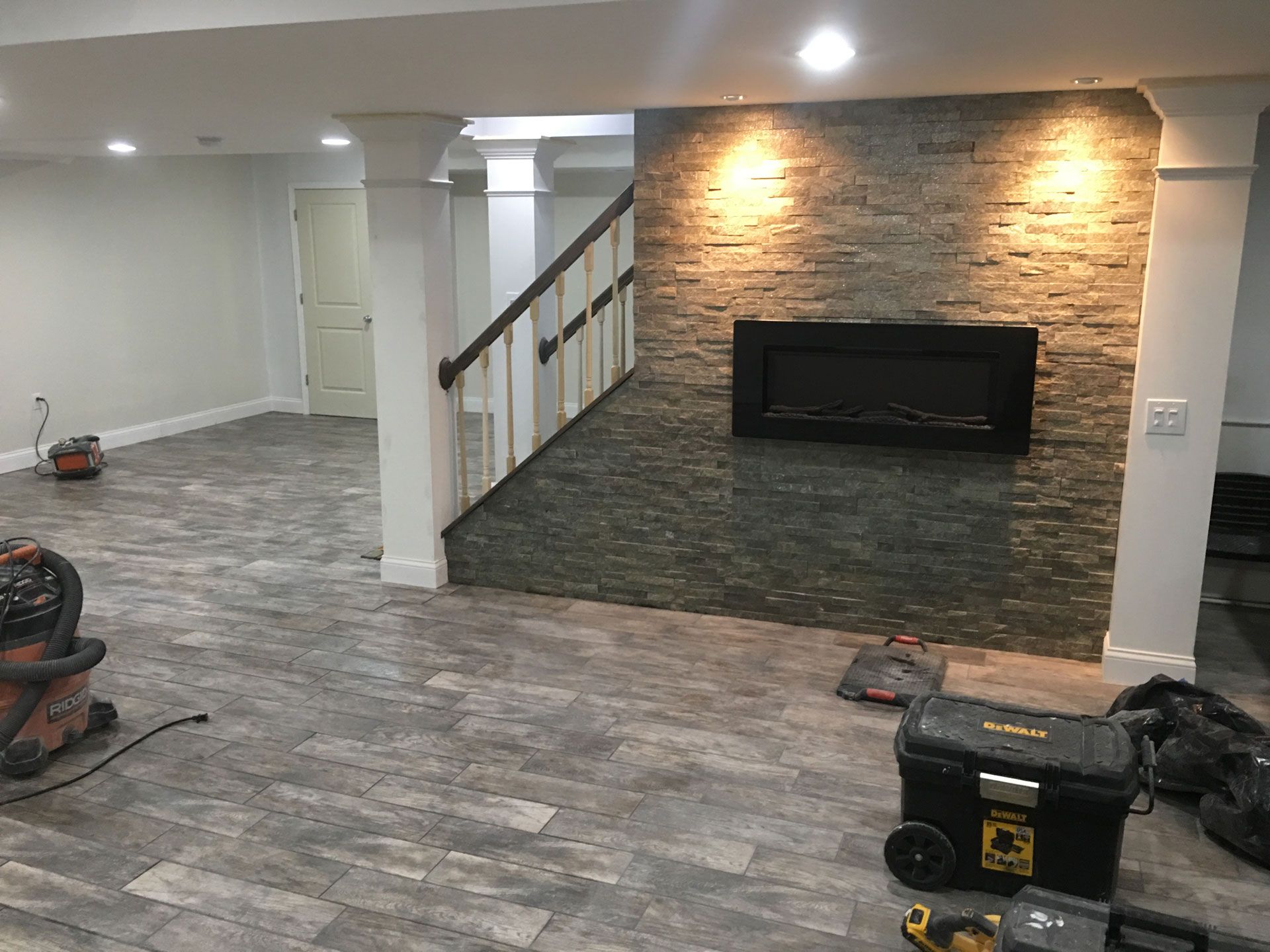 A basement renovation features grey wood-look floors, white support columns, stairs, and a stone wall with a fireplace.