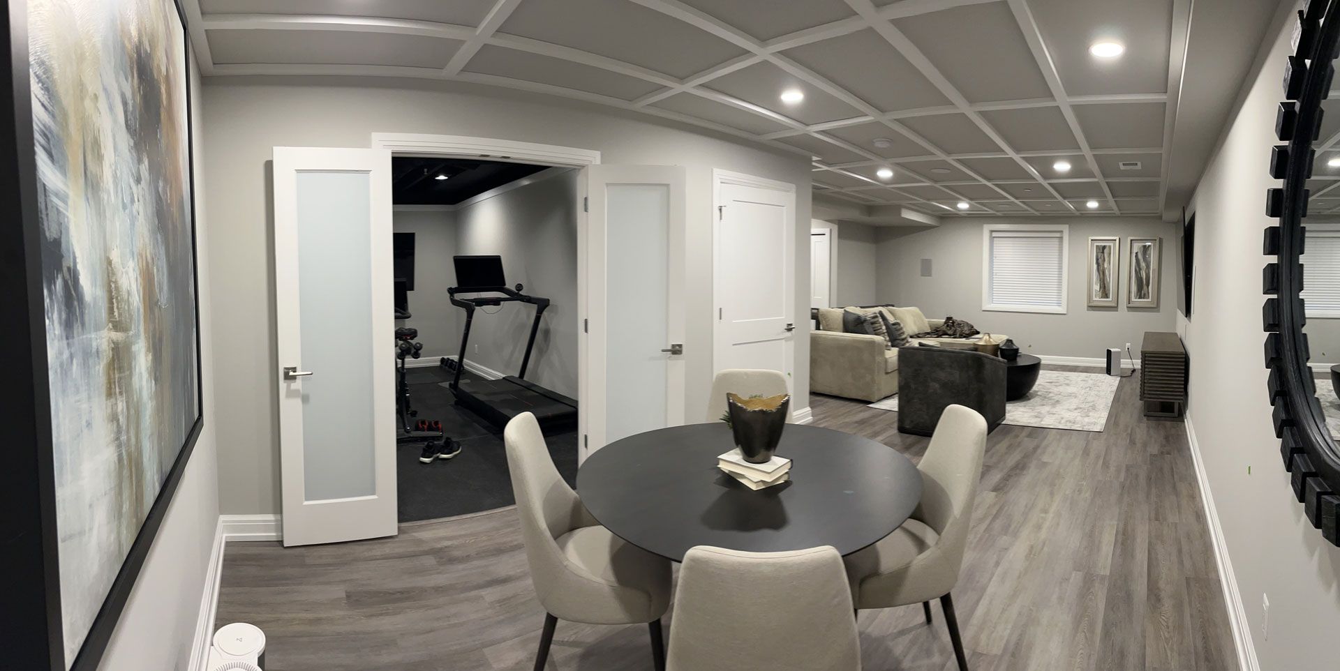 A modern, neutral-toned basement with a dining table, a fitness area with a treadmill, and a cozy lounge space.