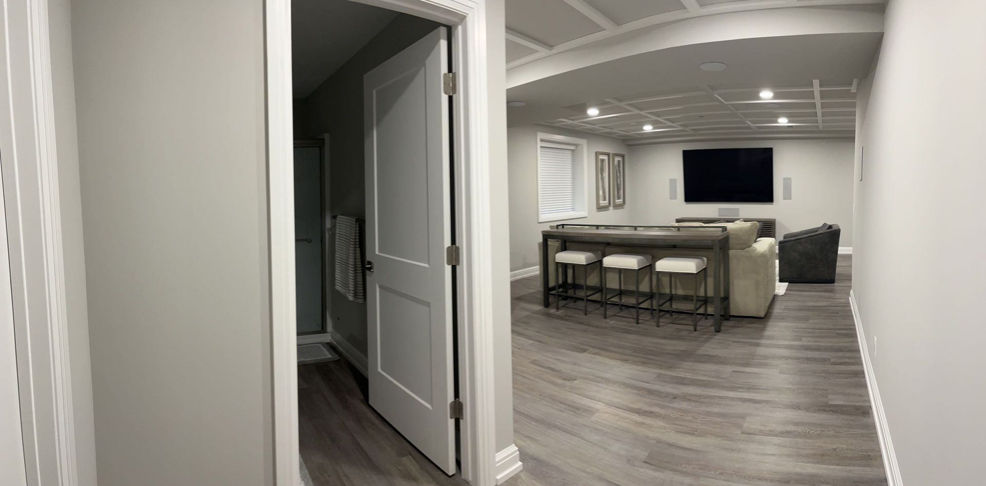 A hallway with an open door to a bathroom, leading into a finished basement living area with seating and a large TV.