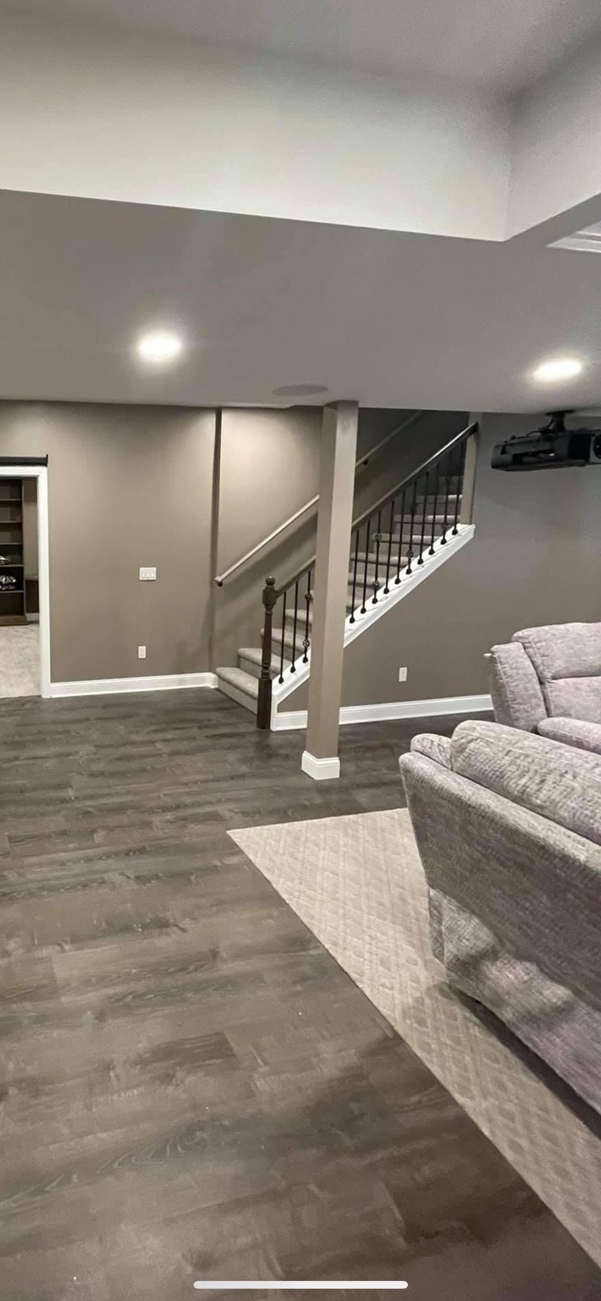 A carpeted staircase with dark railings sits beside a neutral-toned room with wood-look flooring and gray sectional sofa.