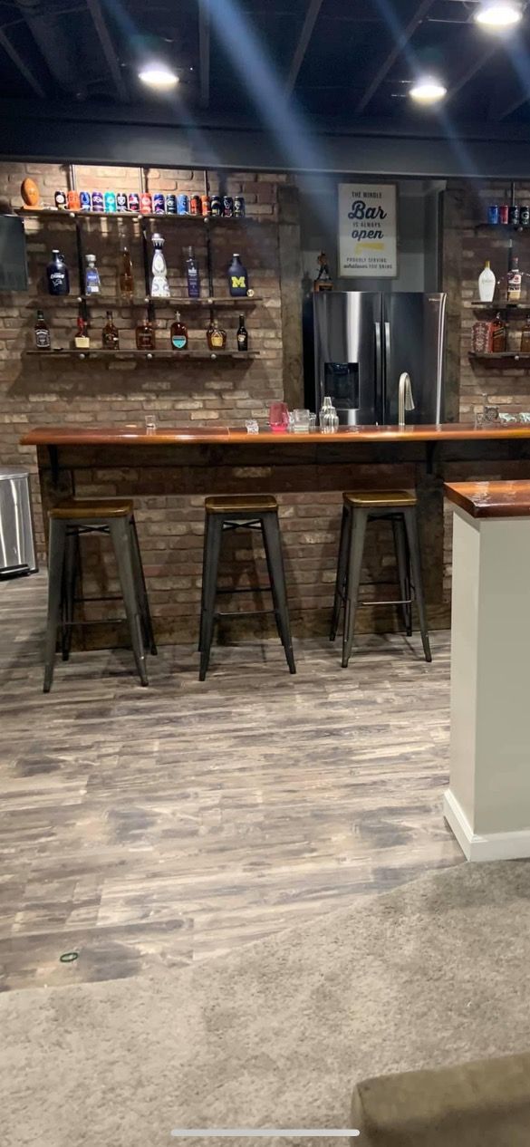 A home basement bar with rustic brick walls, three metal bar stools, wood countertops, and liquor shelves.