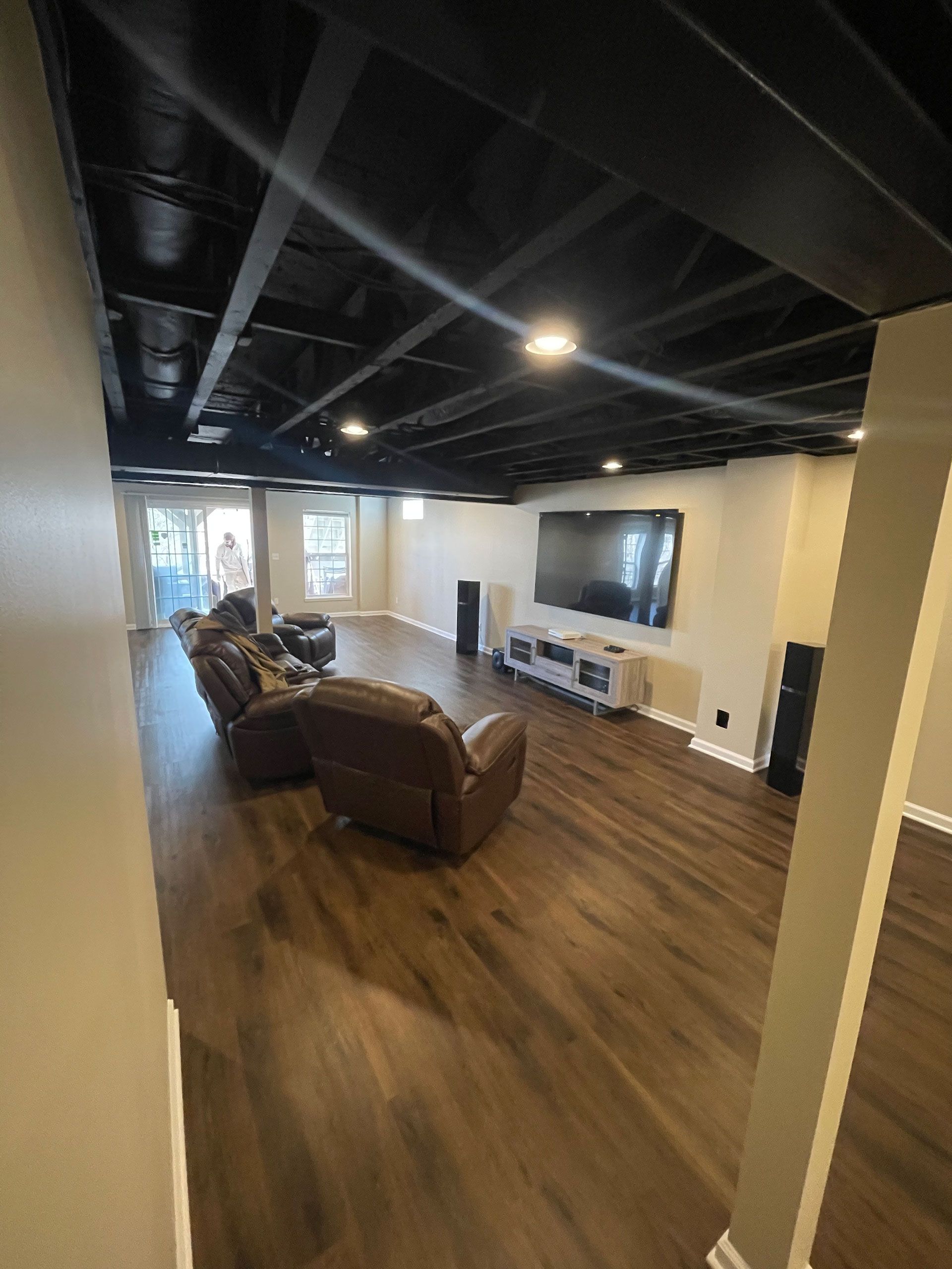 A basement living room with dark exposed joist ceilings, light wood floors, a wall-mounted TV, and brown leather recliners.
