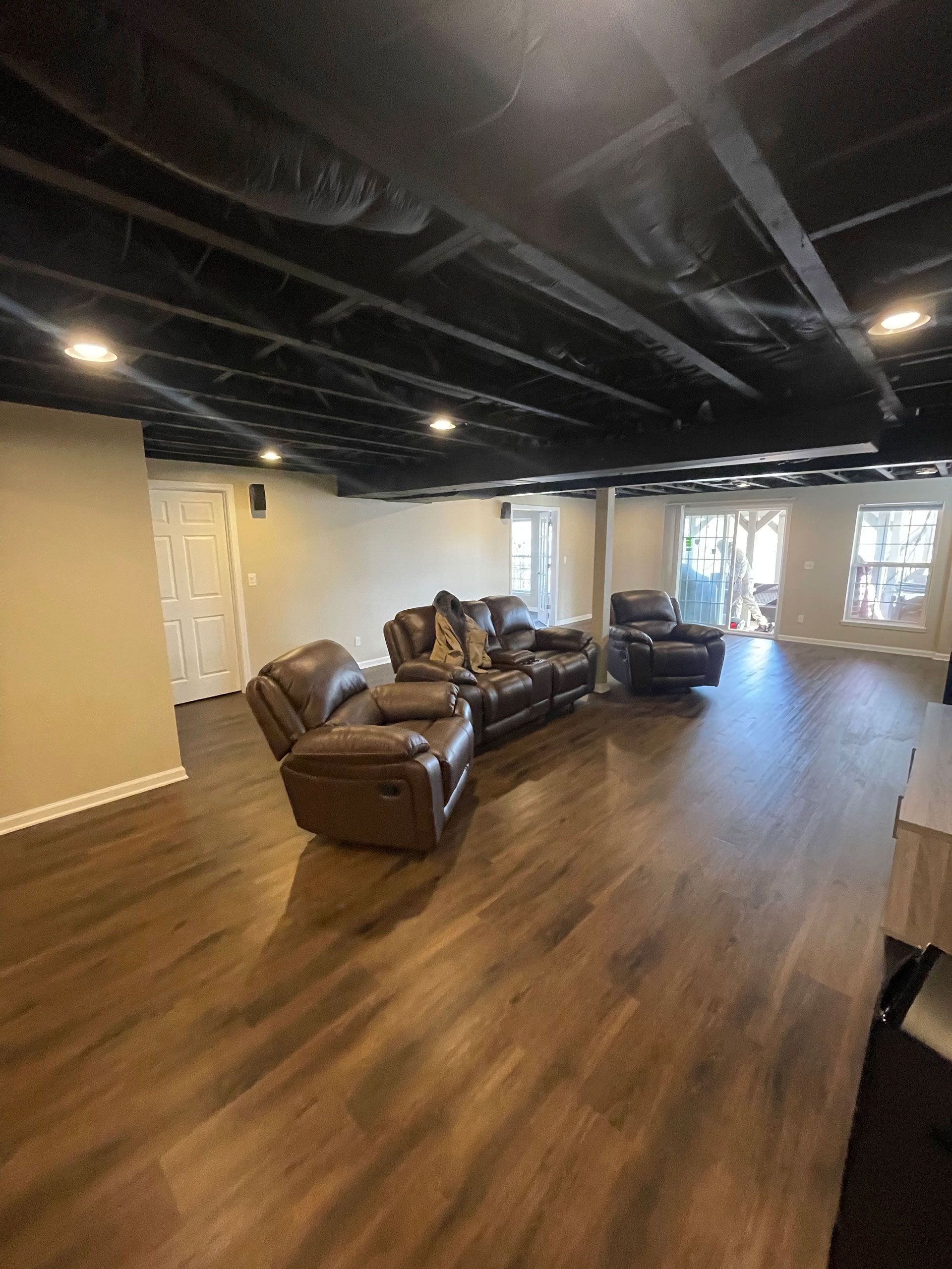 A spacious basement living area with wood-look floors, an open ceiling painted black, and brown leather seating.
