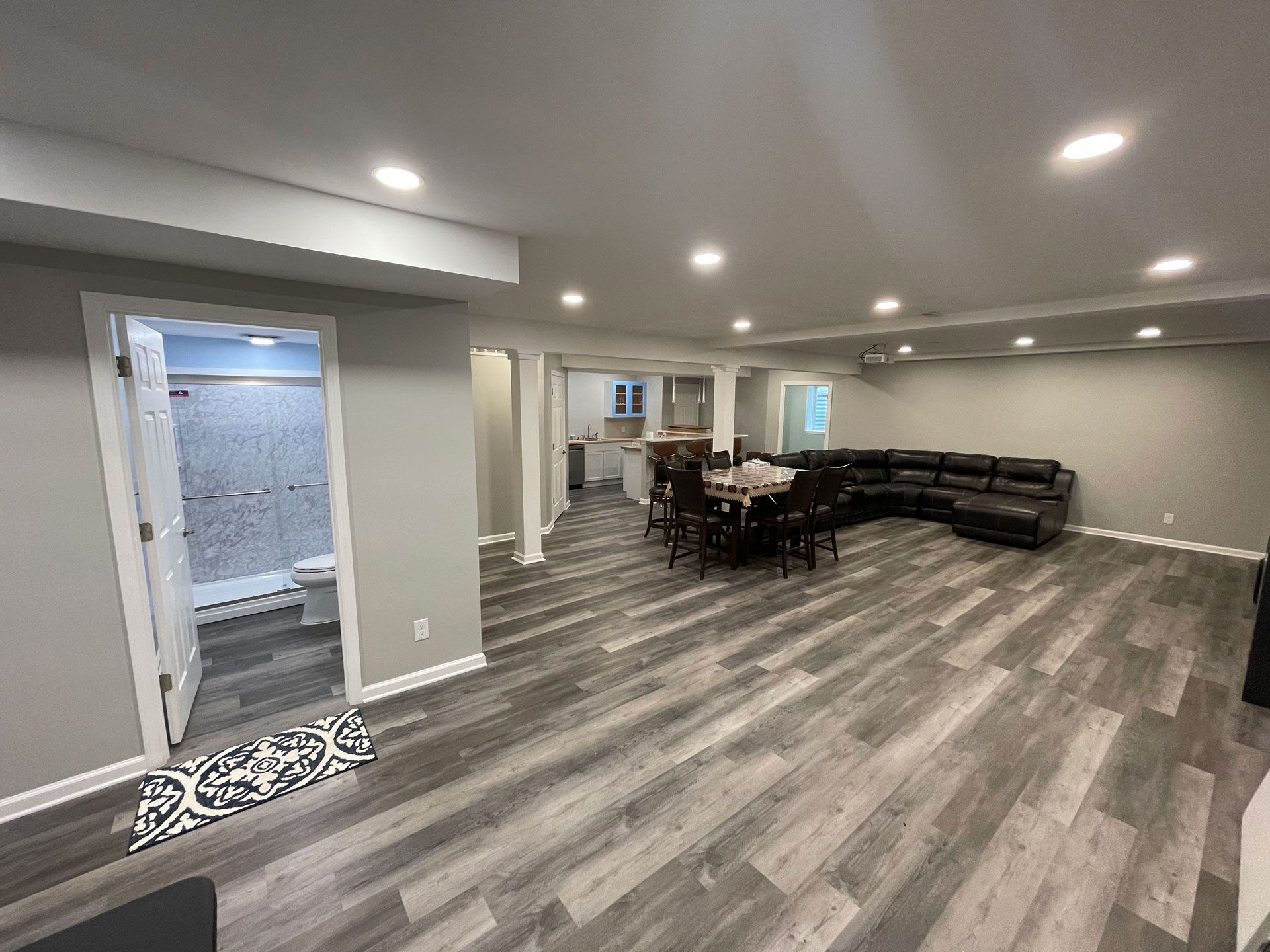 A wide-angle view of a finished basement with grey wood-style flooring, a recessed bathroom, a dining table, and a sofa.