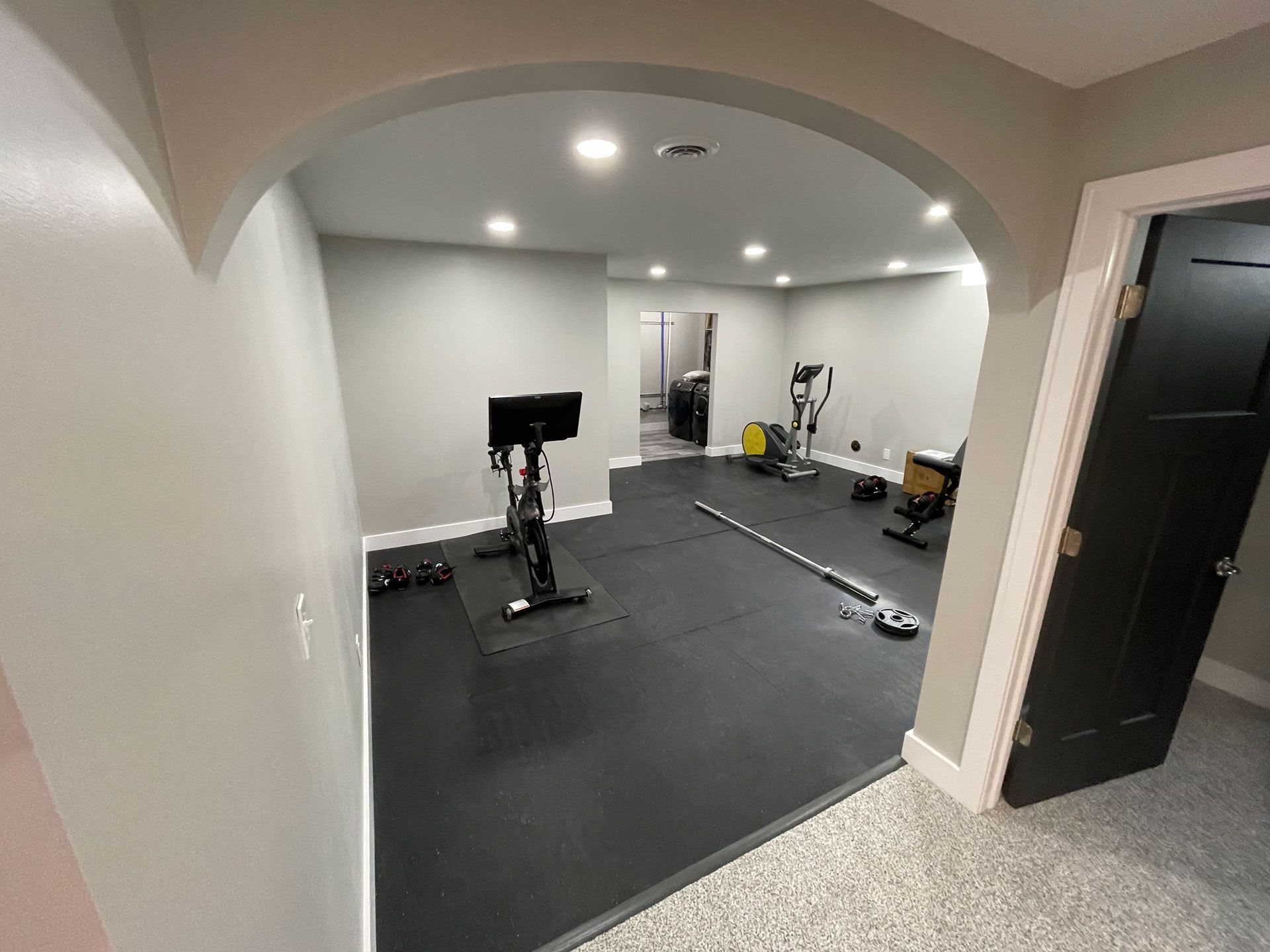 A home gym space with black rubber flooring, a stationary bike, elliptical, and workout bench under an arched entryway.