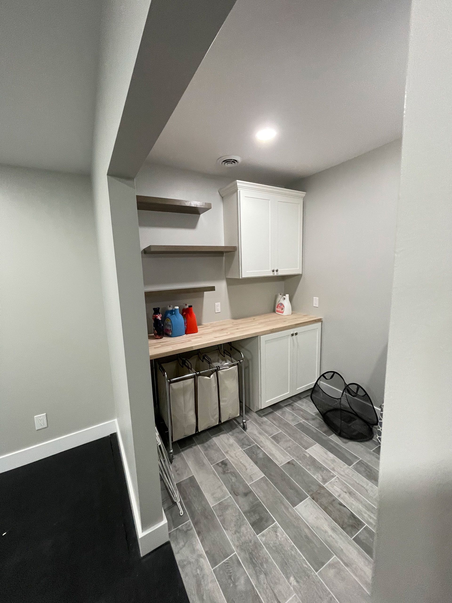 A laundry area featuring wood shelving, white cabinets, a light wood countertop, a rolling hamper, and gray tile flooring.