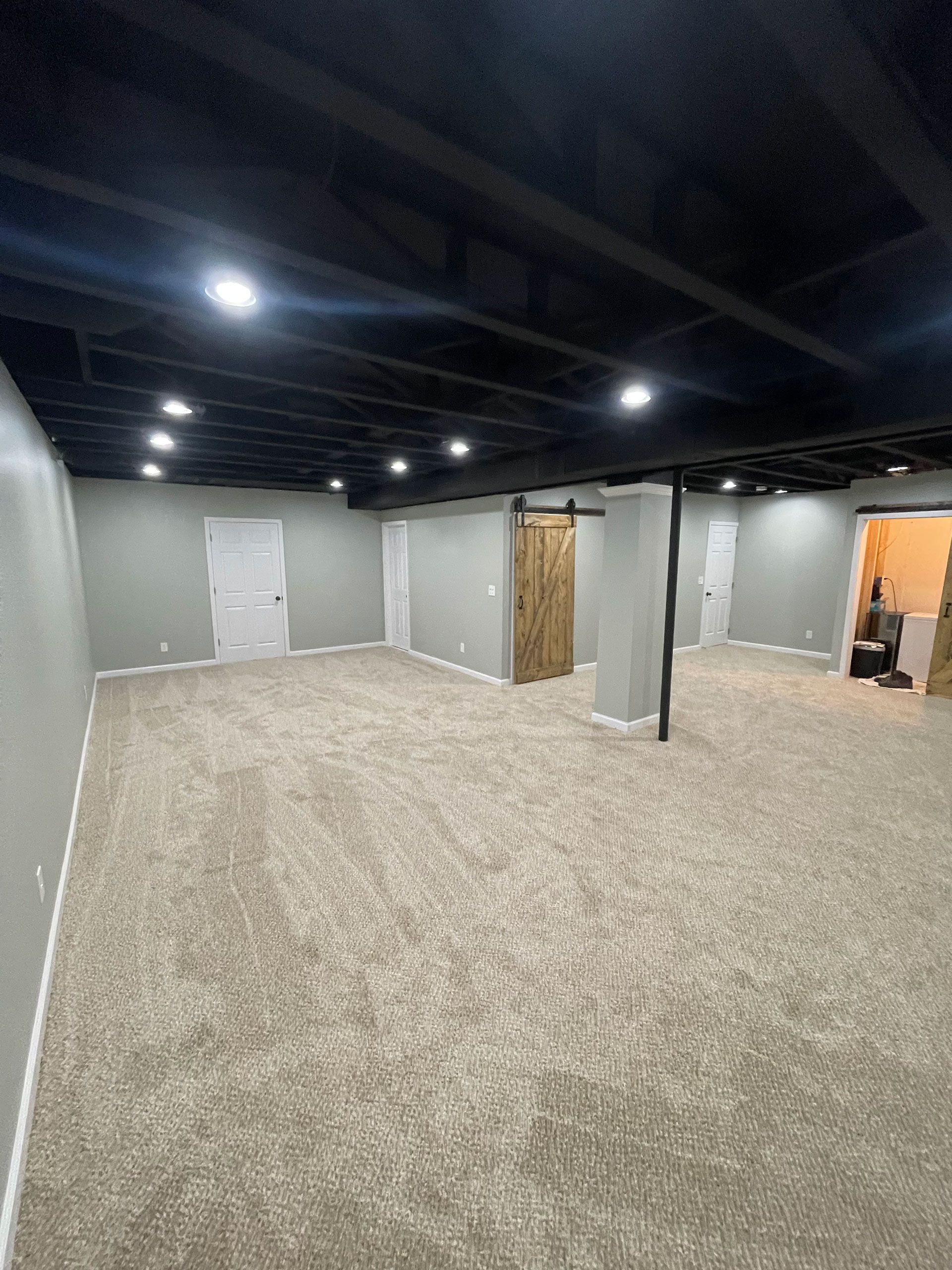 A basement with gray walls, tan carpet, and an exposed black-painted ceiling with recessed lighting.