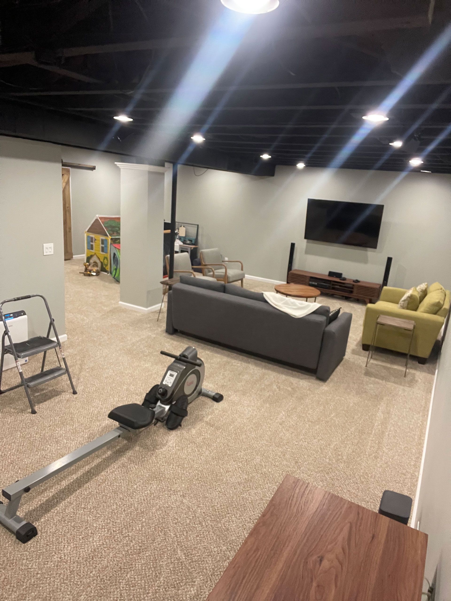 A basement living area with a grey couch, a green armchair, a wall-mounted TV, a rowing machine, and a children's play area.