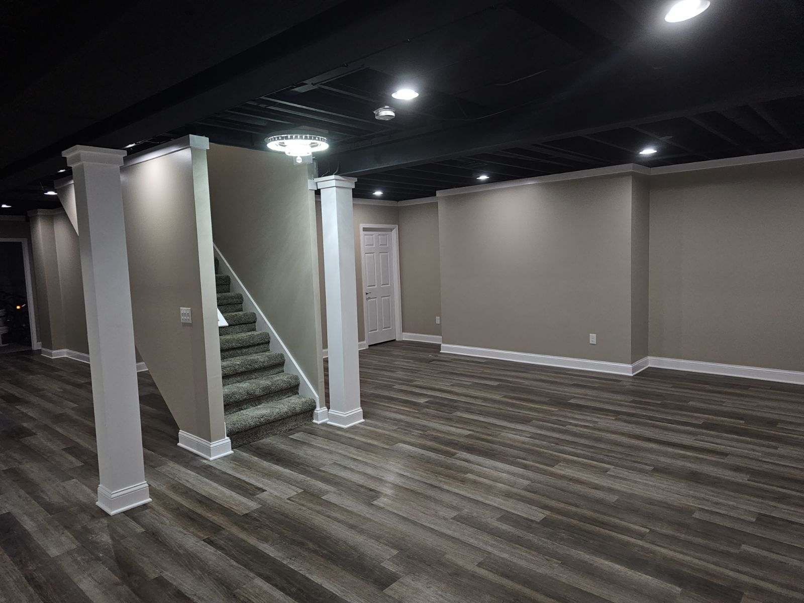 A basement with wood-look flooring, taupe walls, and a black exposed-joist ceiling with recessed lighting and columns.