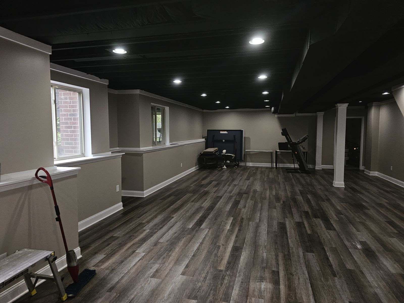 A basement interior featuring grey walls, dark wood-look plank flooring, a black painted ceiling, and recessed lighting.