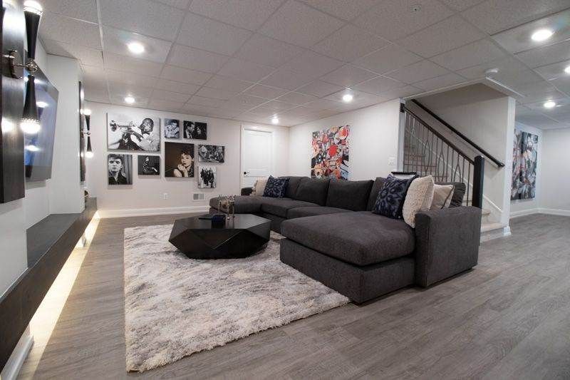 A basement living room features a grey sectional sofa on a plush rug, a coffee table, and wall art under recessed lights.