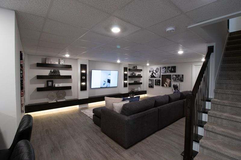 A modern finished basement with a dark gray sofa, wall-mounted television, black shelving, and carpeted stairs.
