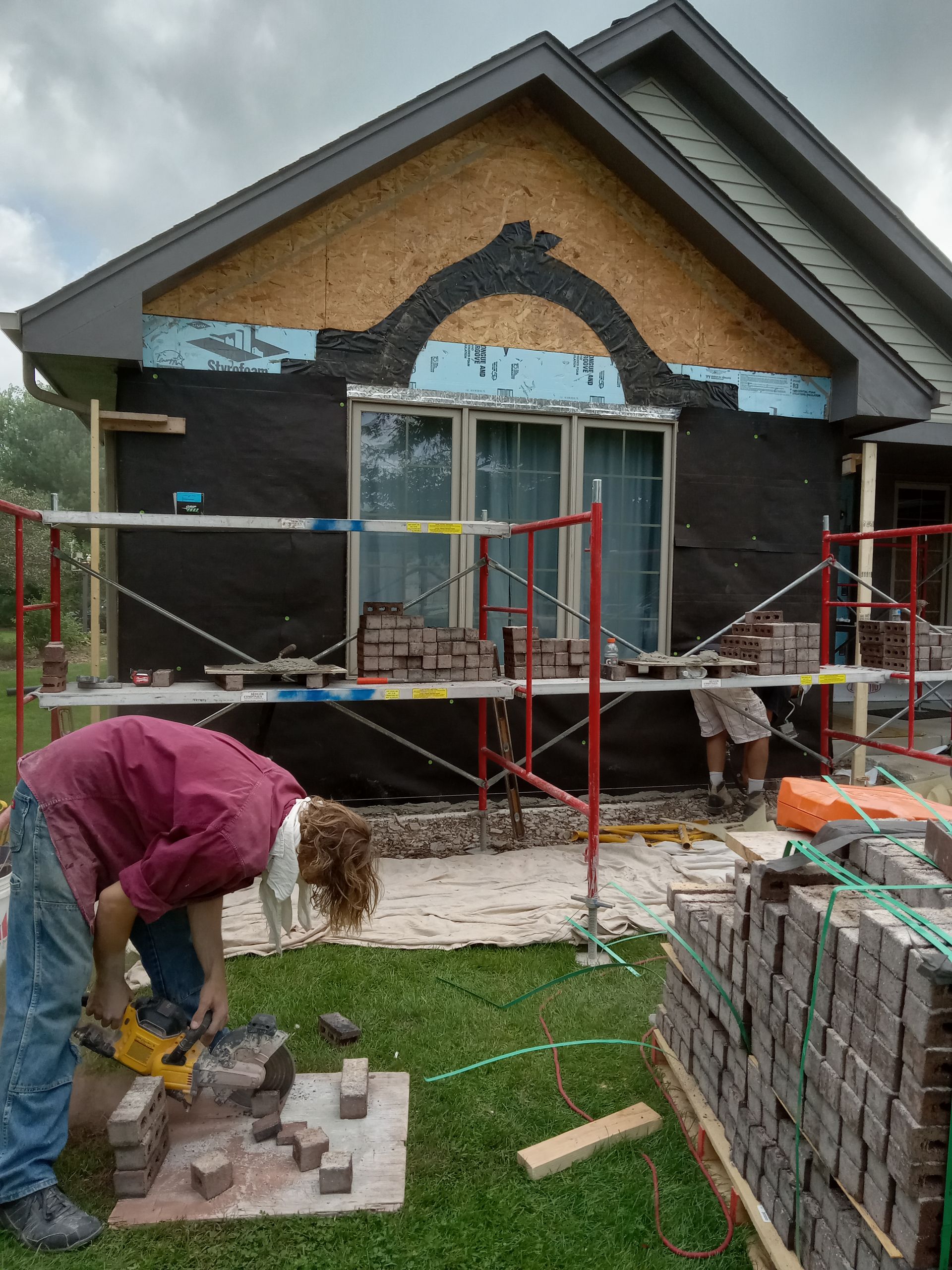 Construction workers laying brick on the side of a house; scaffolding, bricks, and tools are visible.