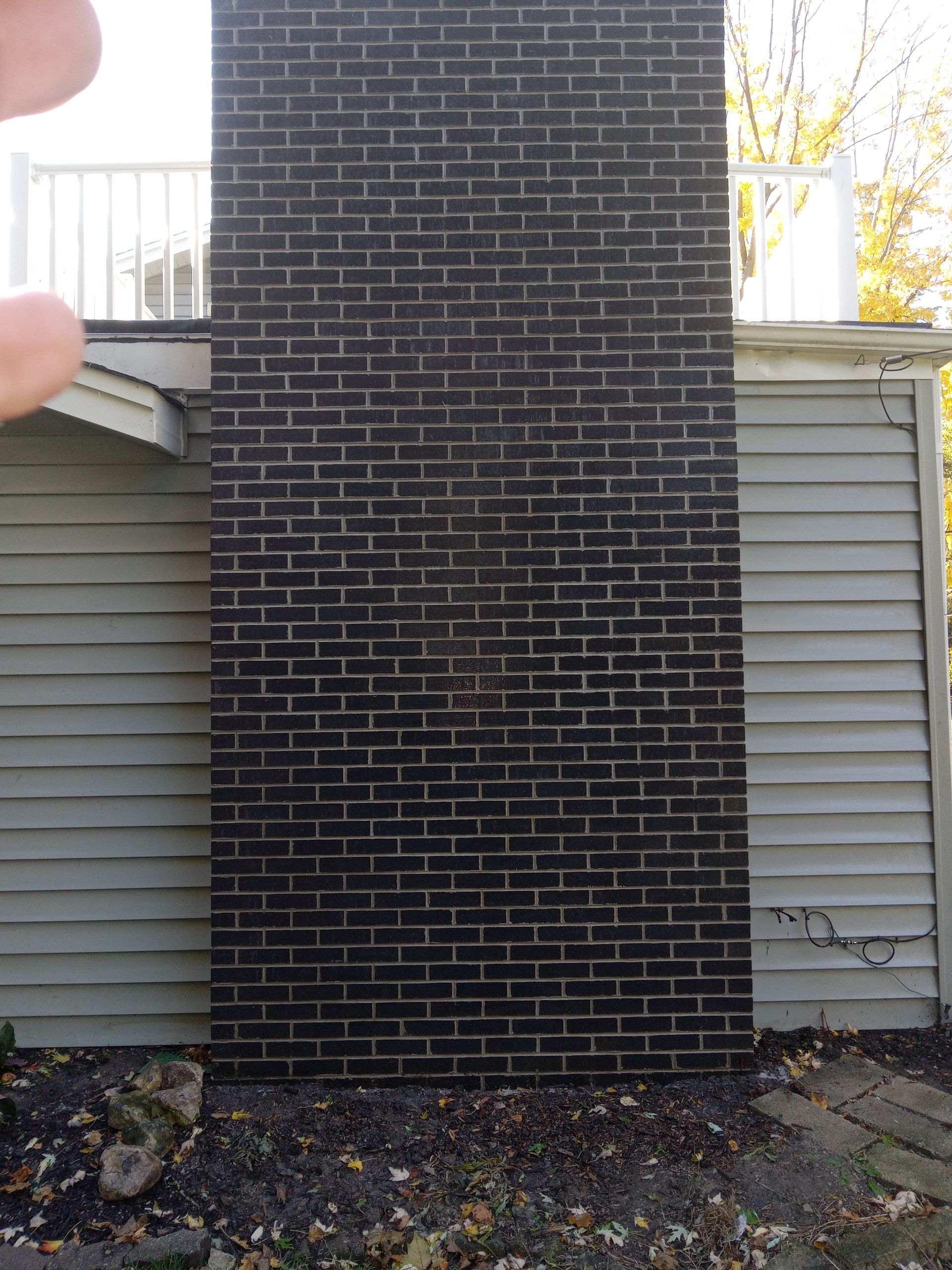 Tall dark brick chimney against a gray house with a white railing.