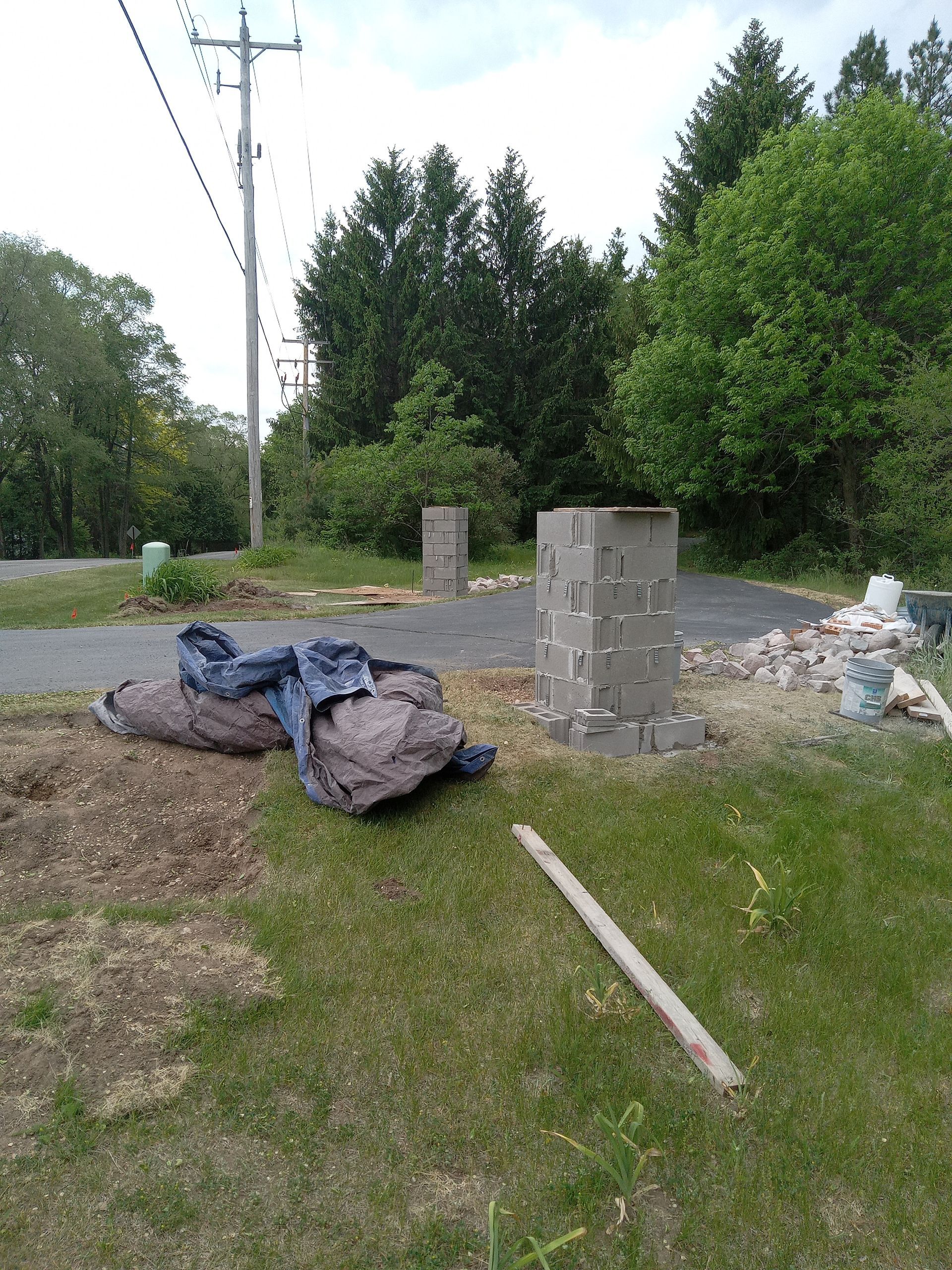 Construction site with three concrete block columns. Gray asphalt road, green grass, and trees.