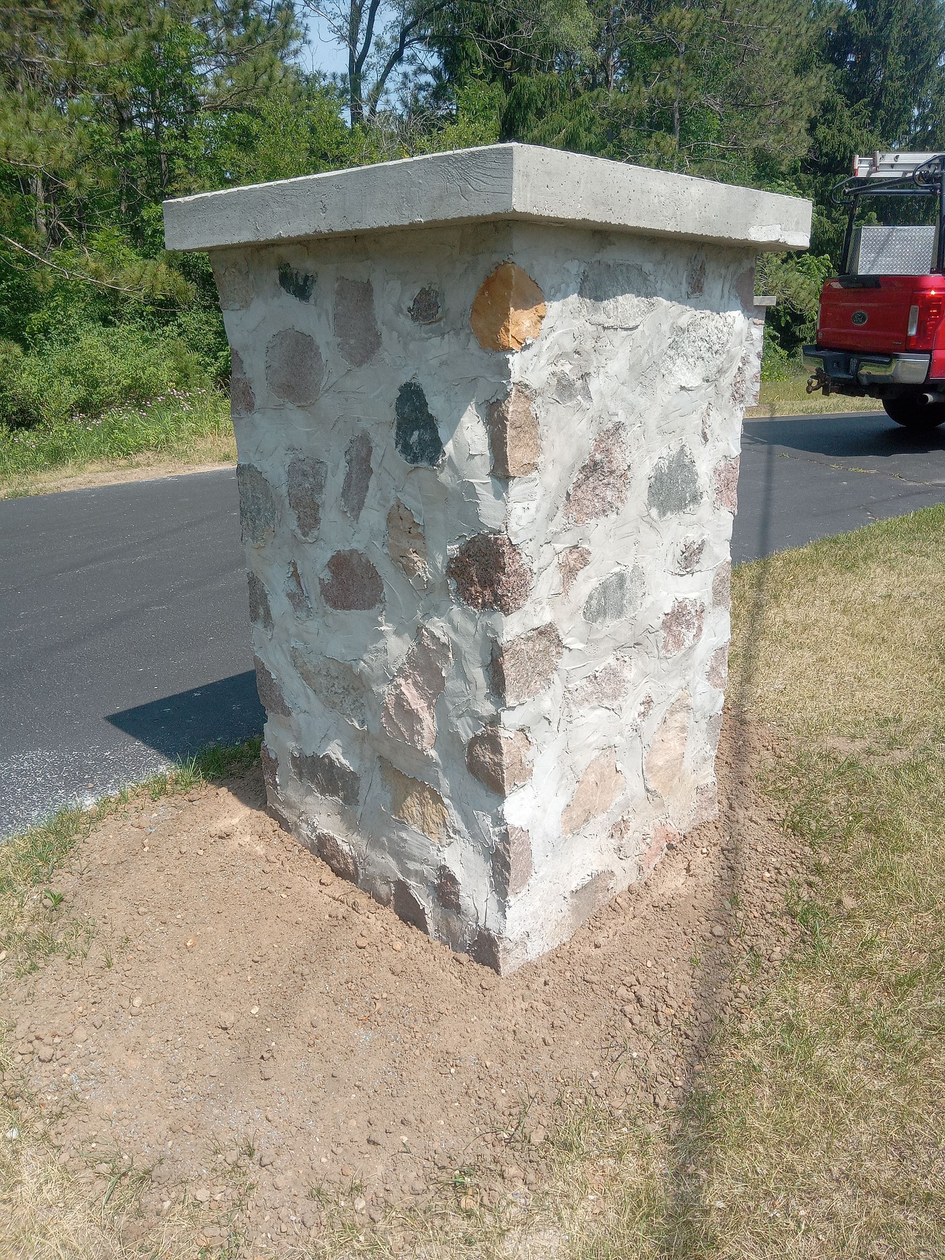 Stone pillar with a concrete cap, next to a road, and a red truck in the background.