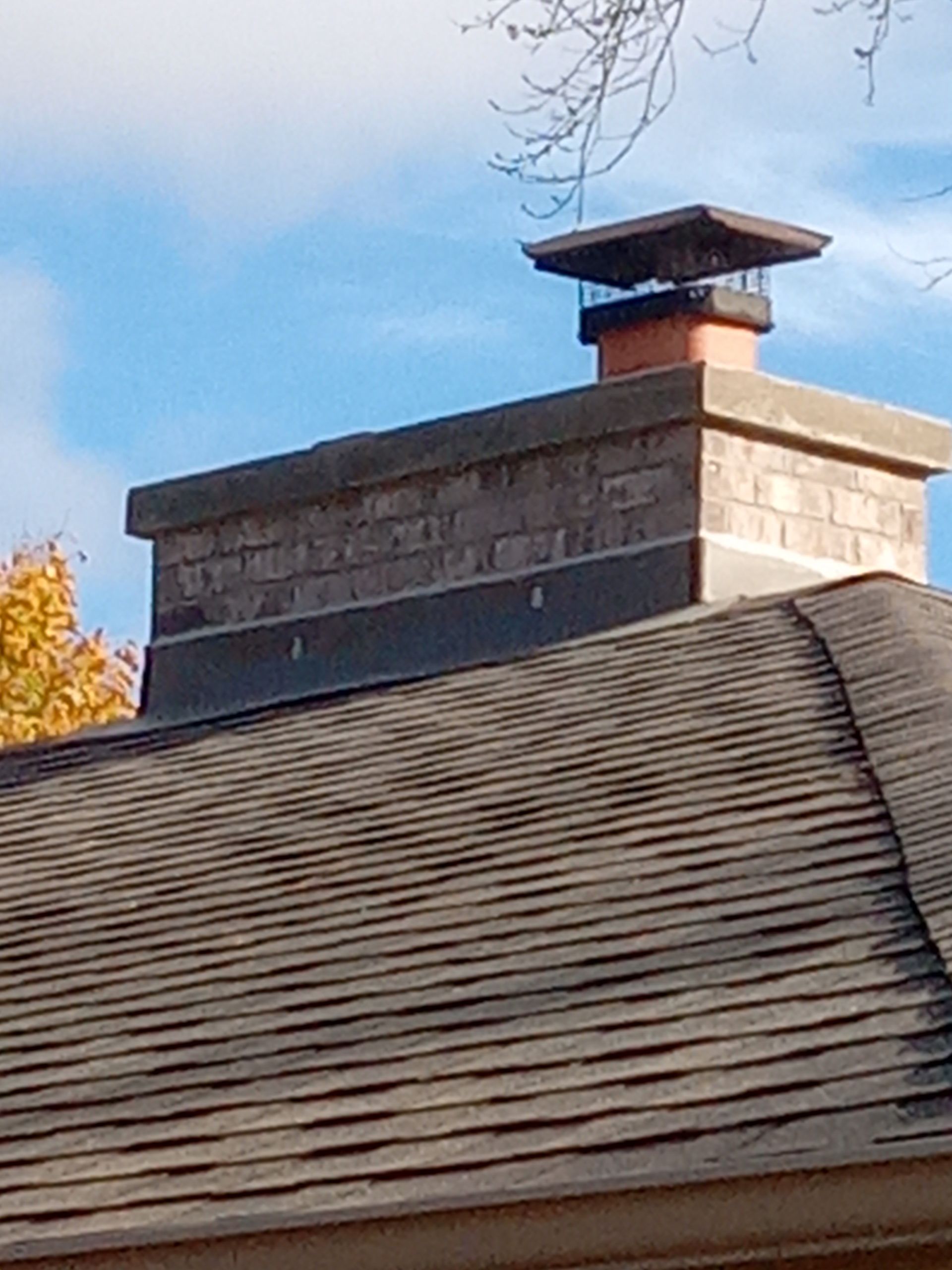 Chimney on a roof, topped with a metal cap. Brown shingles, blue sky.