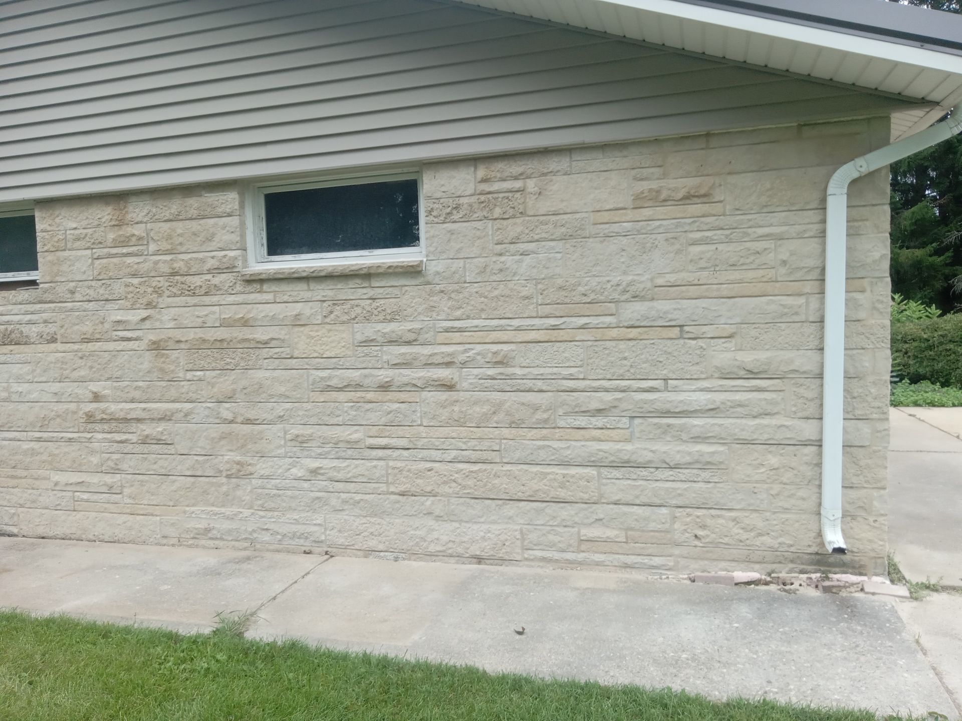 Exterior of a light beige brick wall with a small window, beneath beige siding and a white gutter.