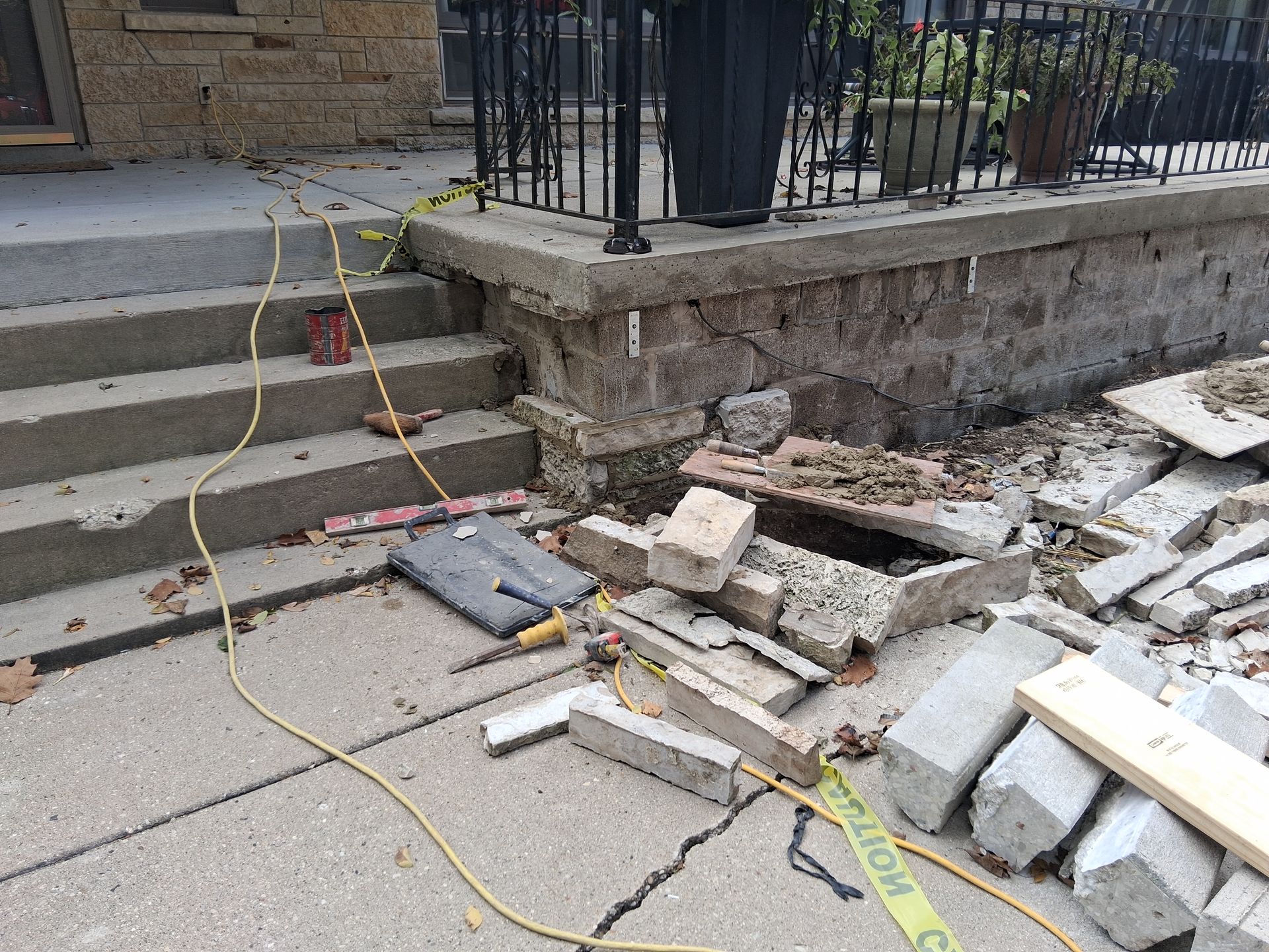 Damaged exterior steps and retaining wall of a building; construction debris on the sidewalk. Yellow electrical cord visible.