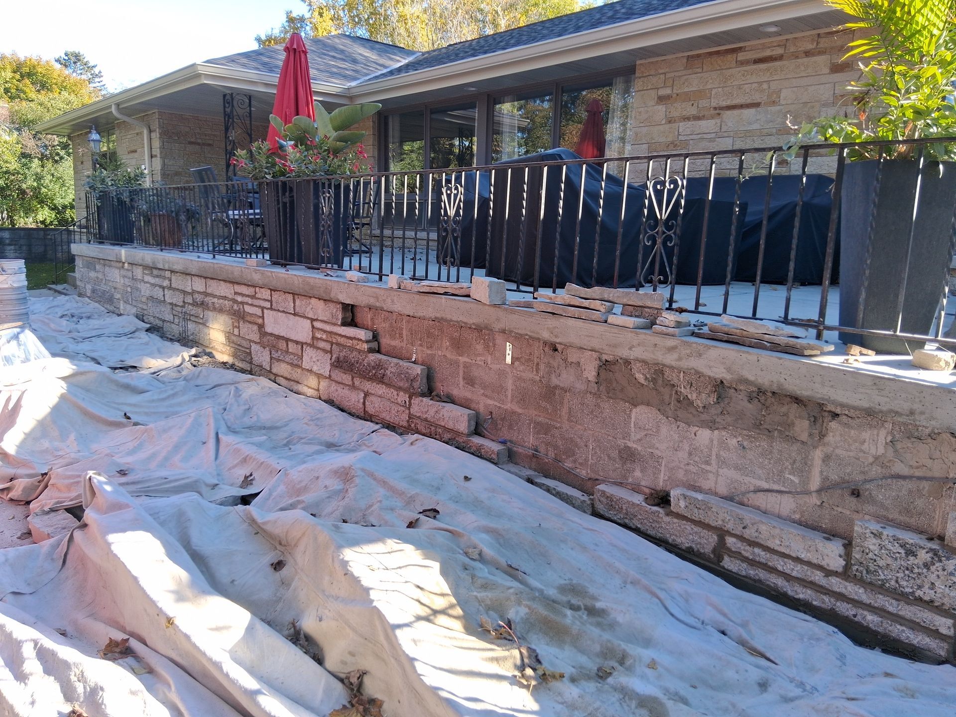 Stone retaining wall under construction next to a house with a black railing and potted plants.