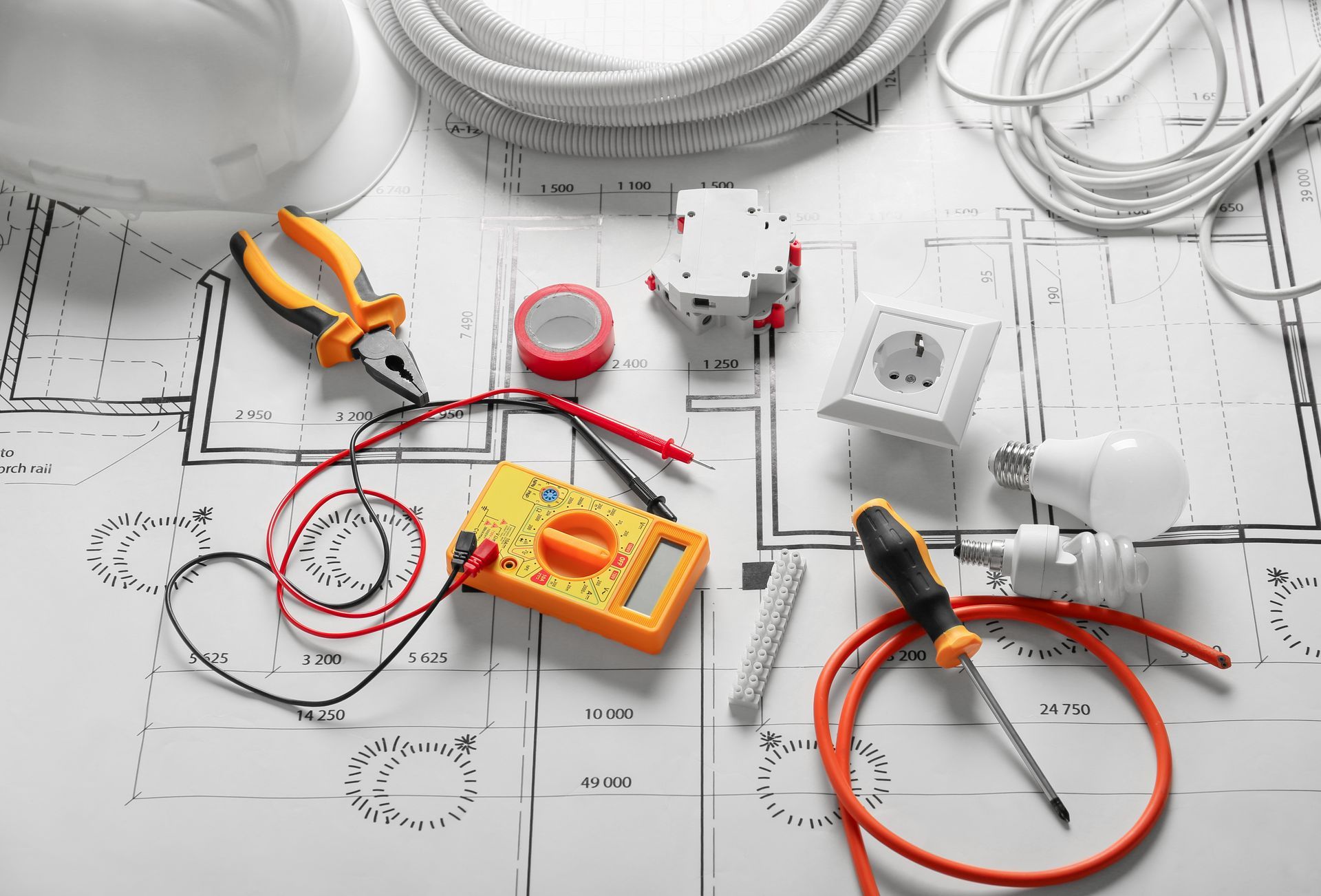 Electrician's tools, wires, and components on a blueprint, including a multimeter and outlet, in a close-up setting.