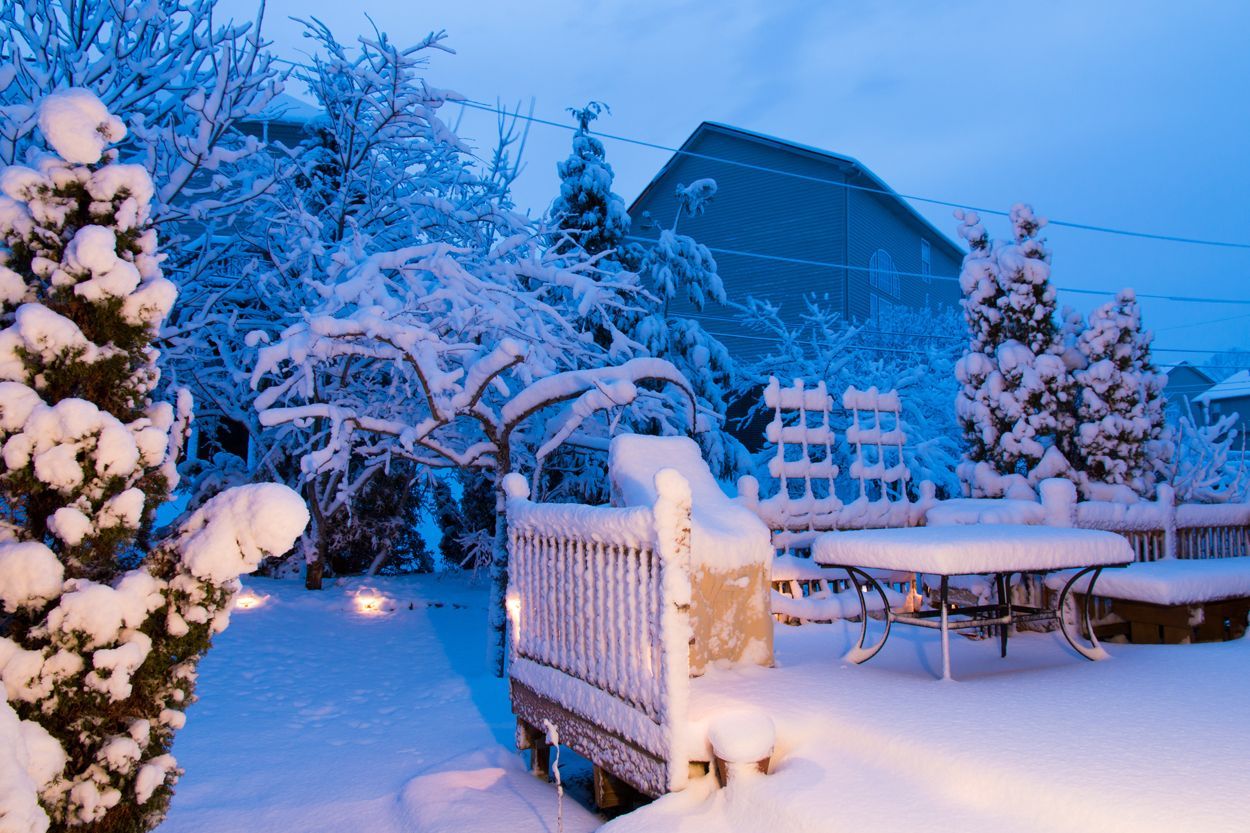 Paysage de jardin enneigé avec mobilier de terrasse et arbres se détachant sur un ciel bleu crépusculaire.