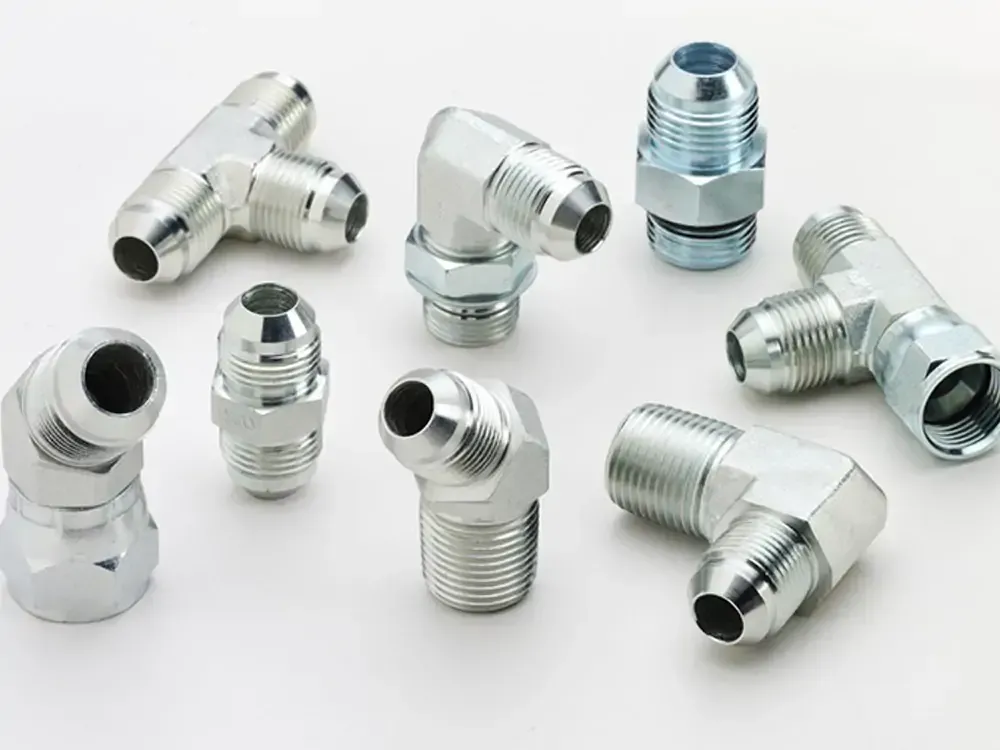 Primer plano de varios accesorios hidráulicos de metal plateado sobre fondo blanco. Los accesorios incluyen codos, tes y adaptadores.