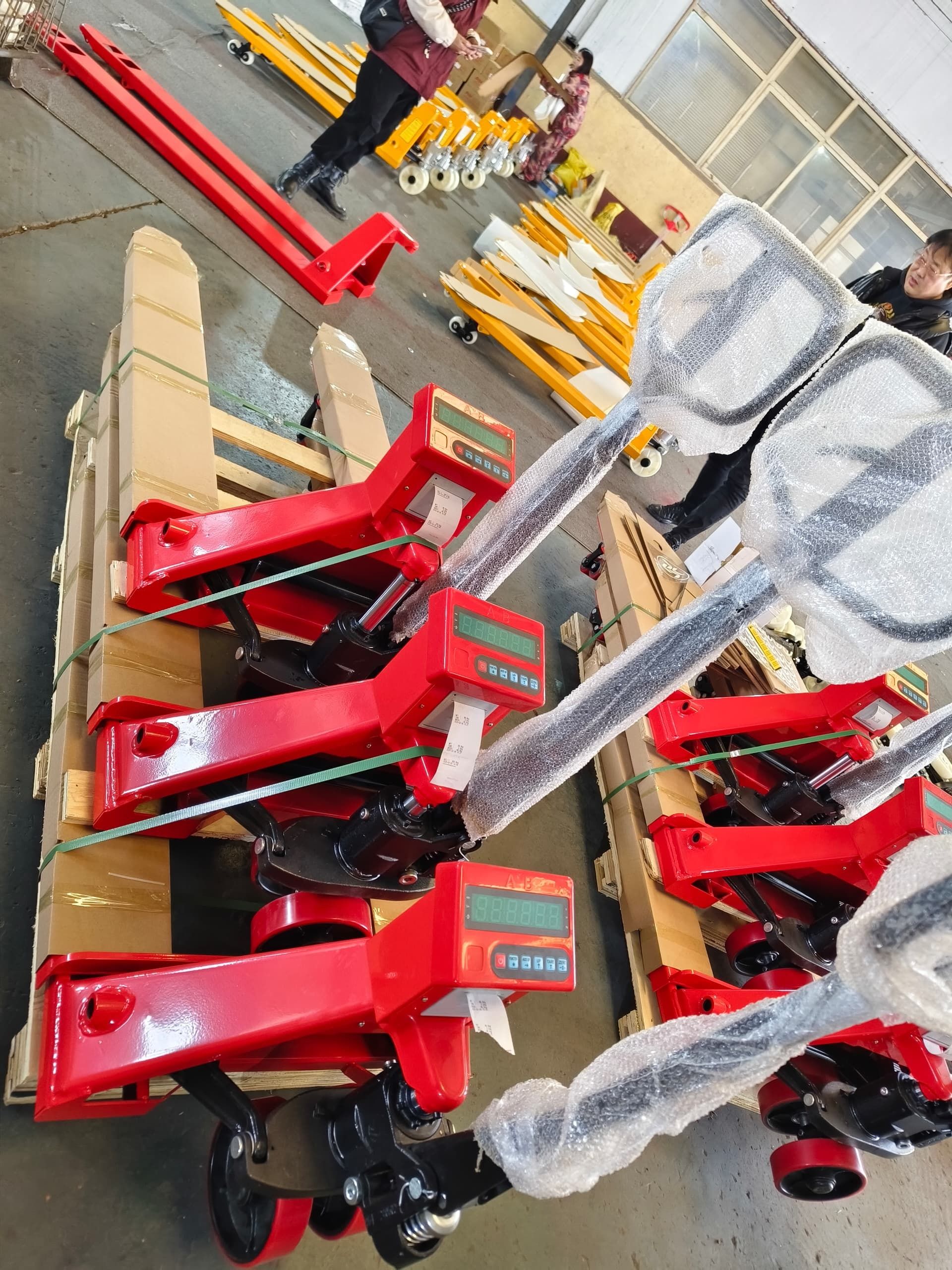 Transpaletas rojas con básculas digitales apiladas sobre paletas de madera en un almacén, con gente en el fondo.