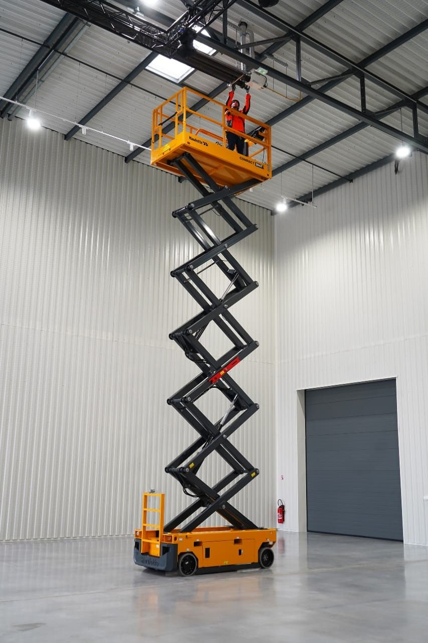 Una persona en una plataforma elevadora de tijera amarilla trabaja en un almacén grande y vacío. La plataforma está extendida, alcanzando el techo.