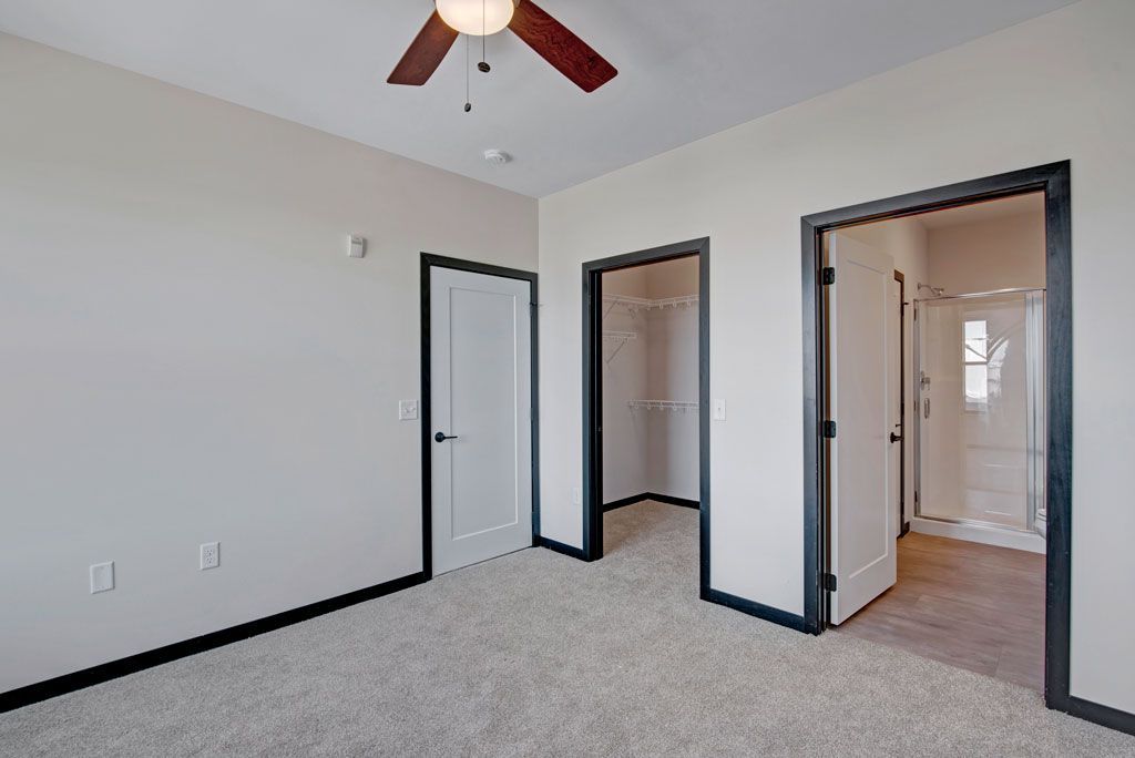 Empty bedroom with white walls, gray carpet, black trim, ceiling fan, and open closet and bathroom doors